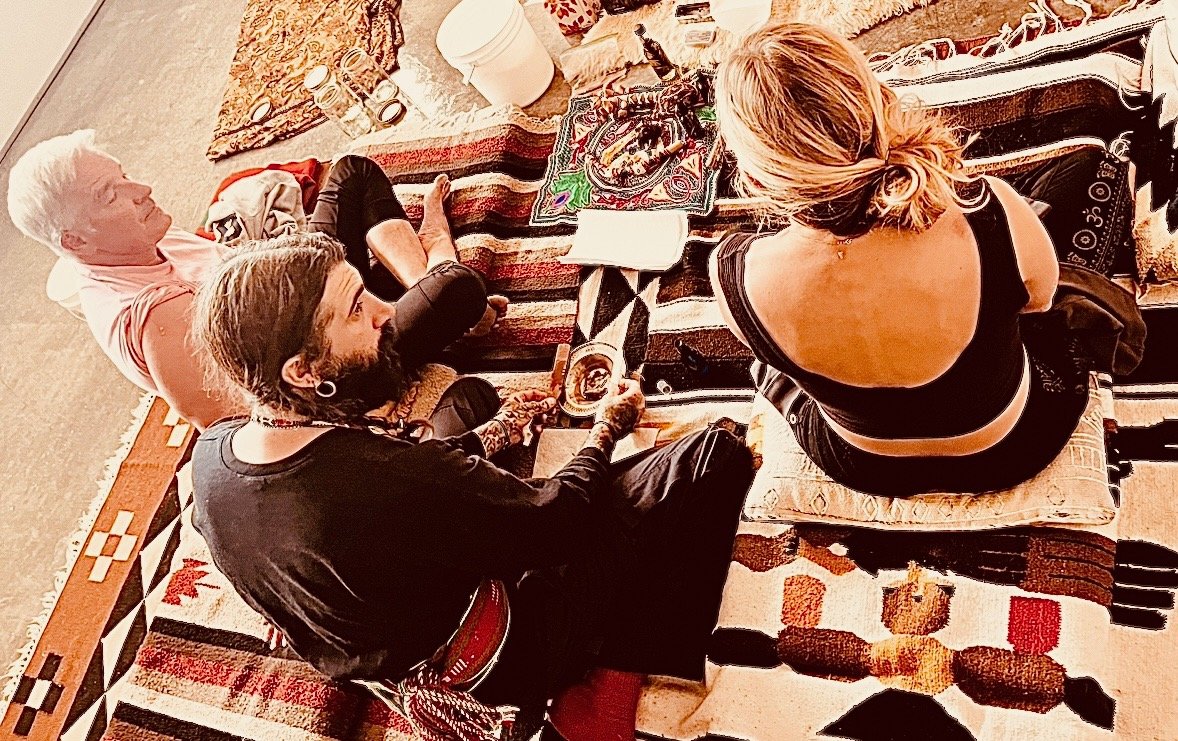 Four people are sitting on cushions on a colorful rug, with a table nearby. They are having a casual gathering, some drinking beverages and sharing food. The scene takes place indoors in a cozy, decorated space.