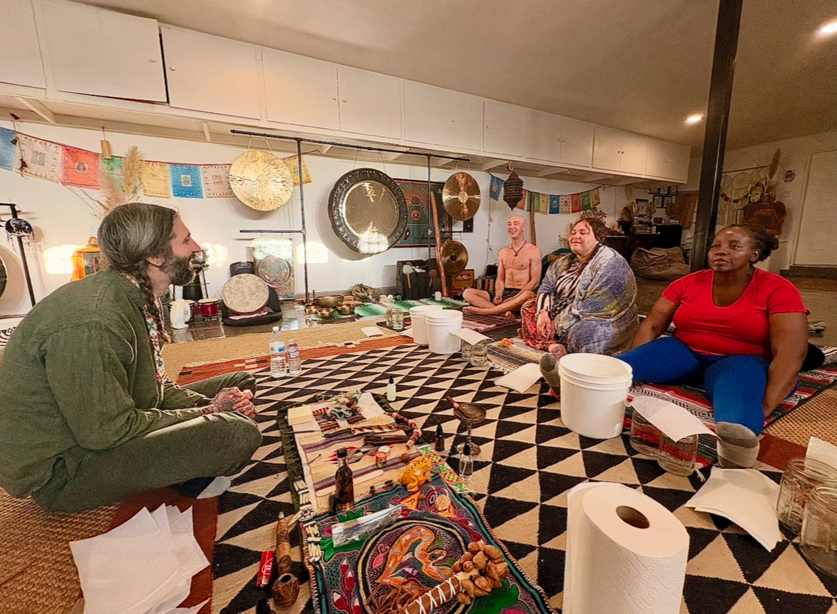 Four people sitting on the floor in a circle in a cozy room decorated with musical instruments, colorful banners, and artwork, participating in a meditation or discussion session.