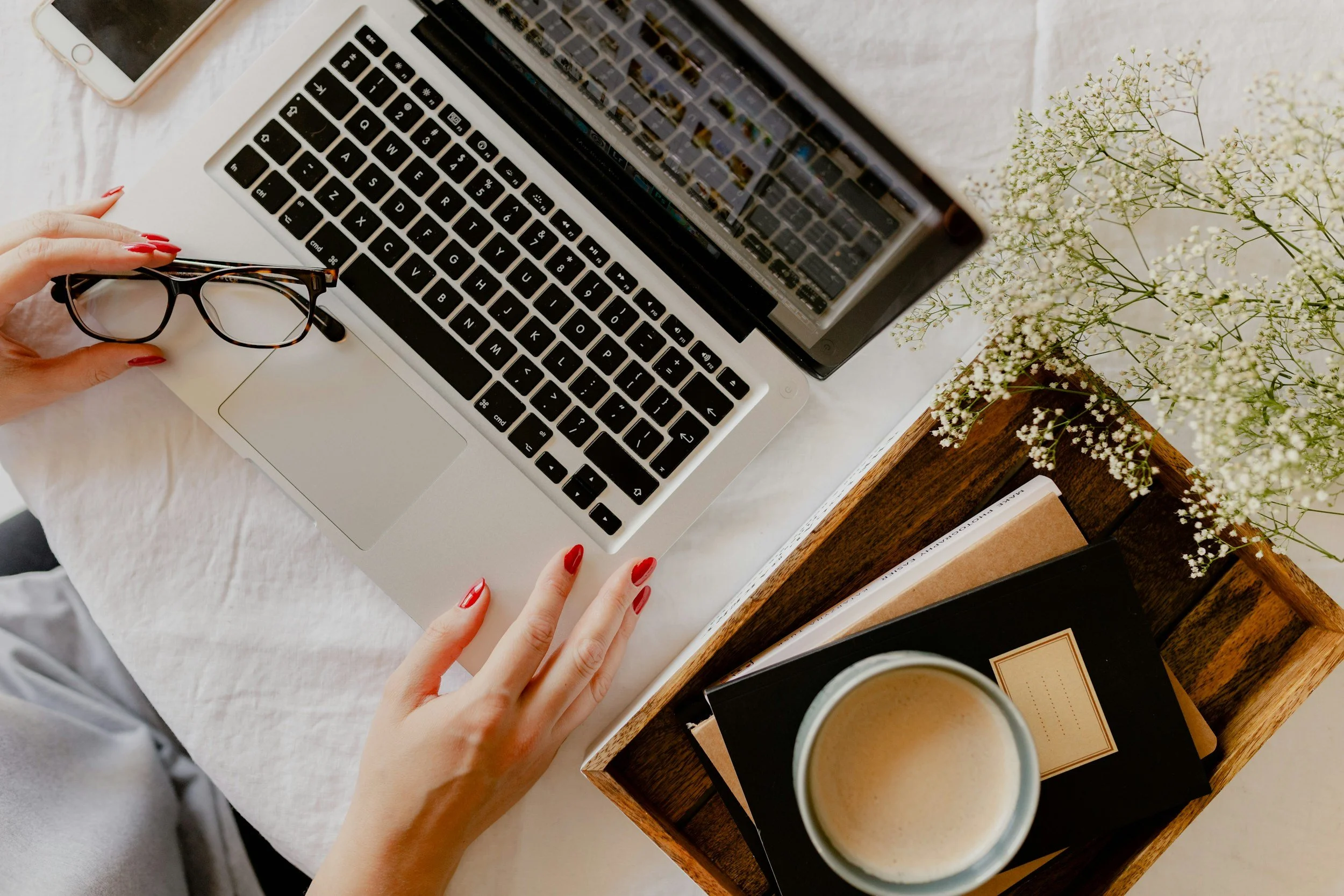 Person with red painted nails using a laptop on a white textured desk, holding glasses in one hand, with a smartphone nearby. There is a wooden tray with notebooks and a cup of coffee, and a vase with white flowers on the desk.