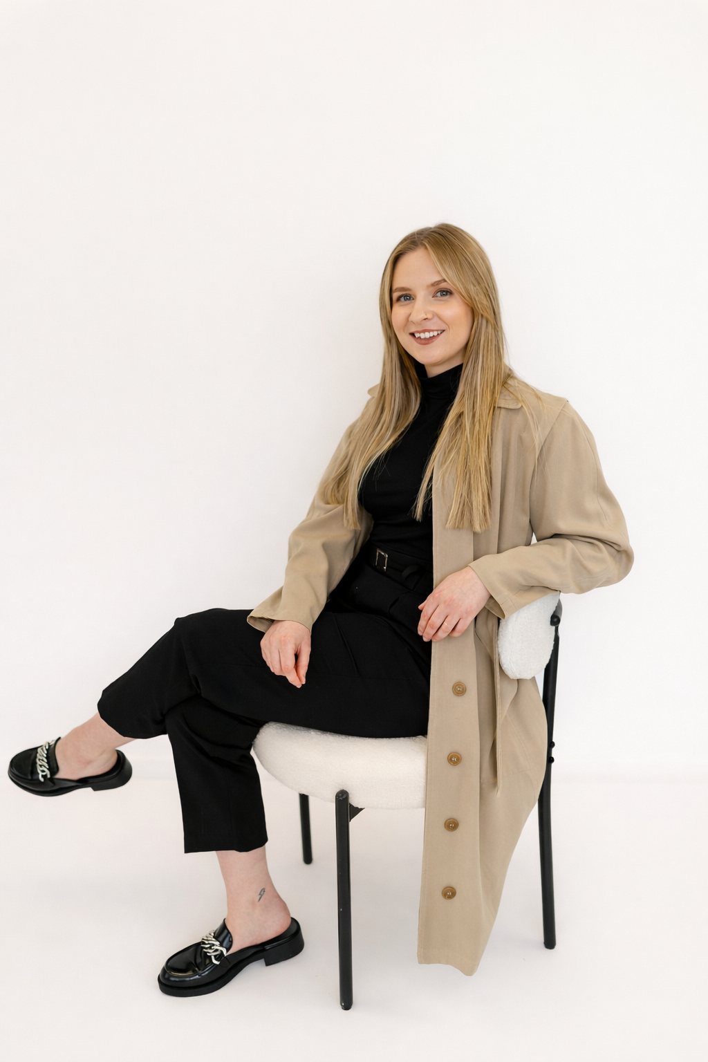 A woman with long blonde hair, wearing a beige trench coat over a black turtleneck, and black pants, sitting on a white chair against a plain white background.