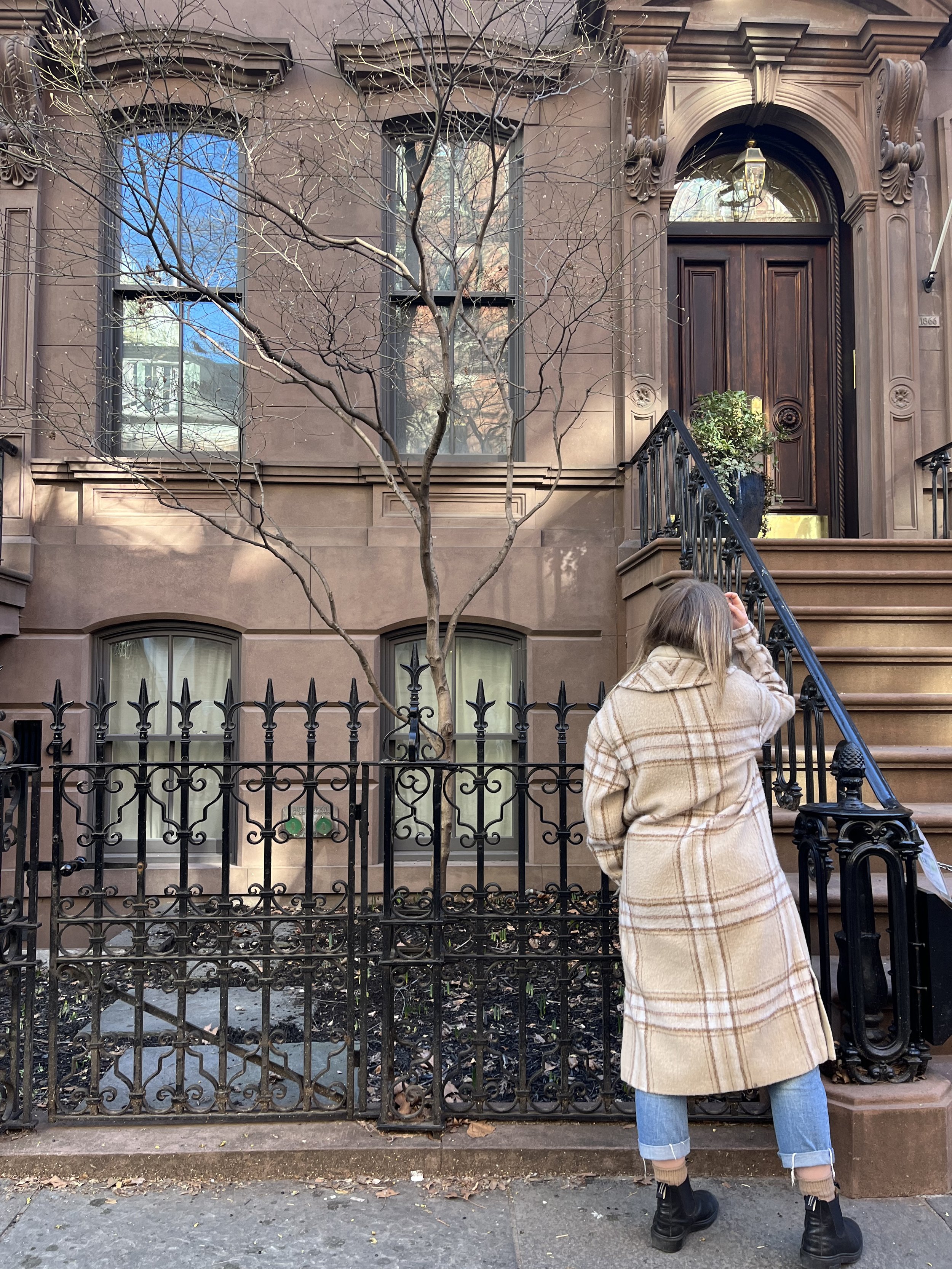 A person standing outside a brownstone building, looking at the door, wearing a long beige plaid coat, jeans, and black boots. The building has a black iron fence and stairs leading up to a wooden door, with a leafless tree in front.
