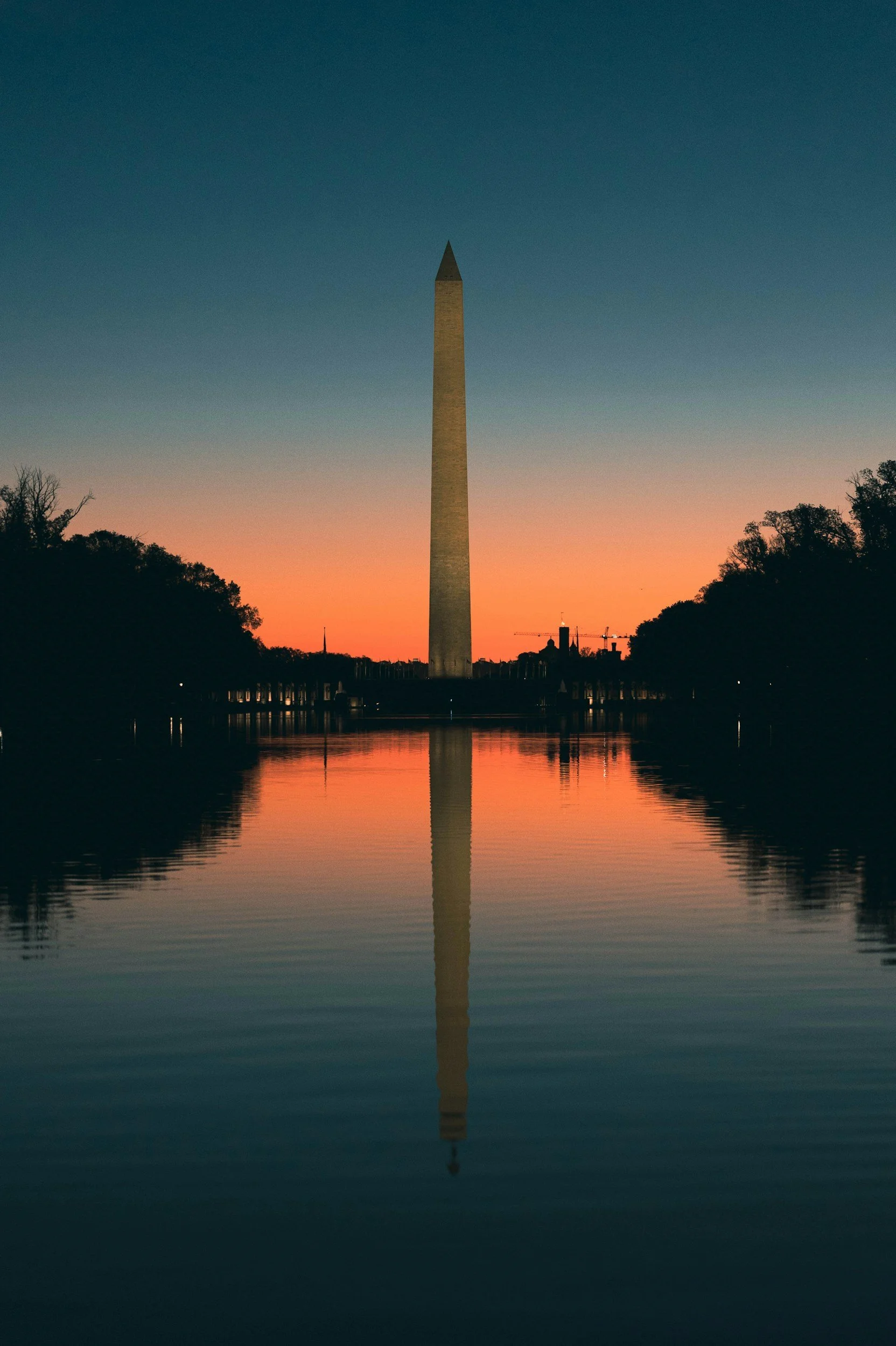 The Washington Monument standing tall with a reflection in the Reflecting Pool at sunset, surrounded by a silhouette of trees against a colorful sky.