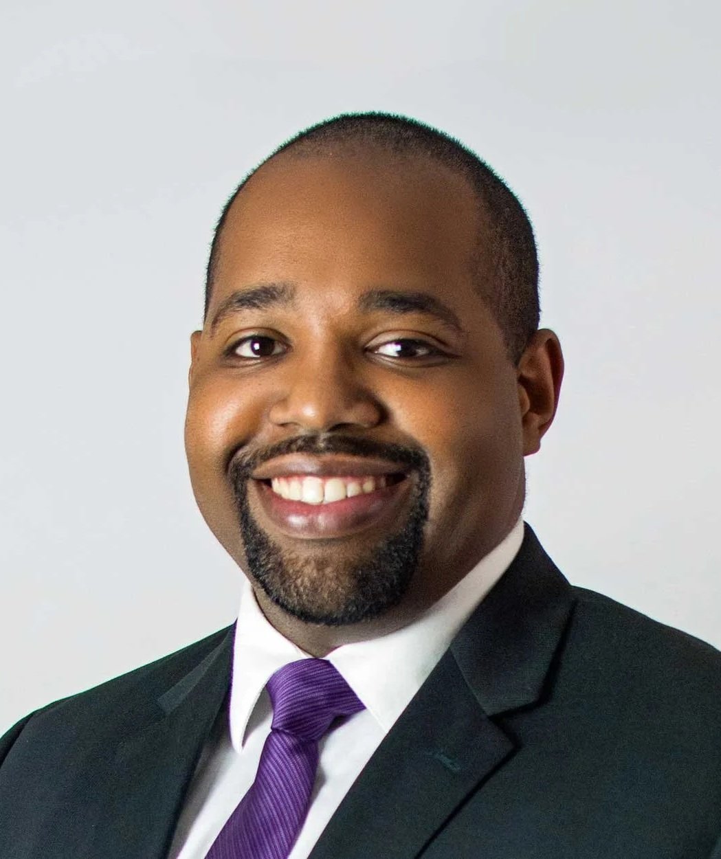 Professional headshot of an African American man in a suit with a purple tie, smiling with a white background.