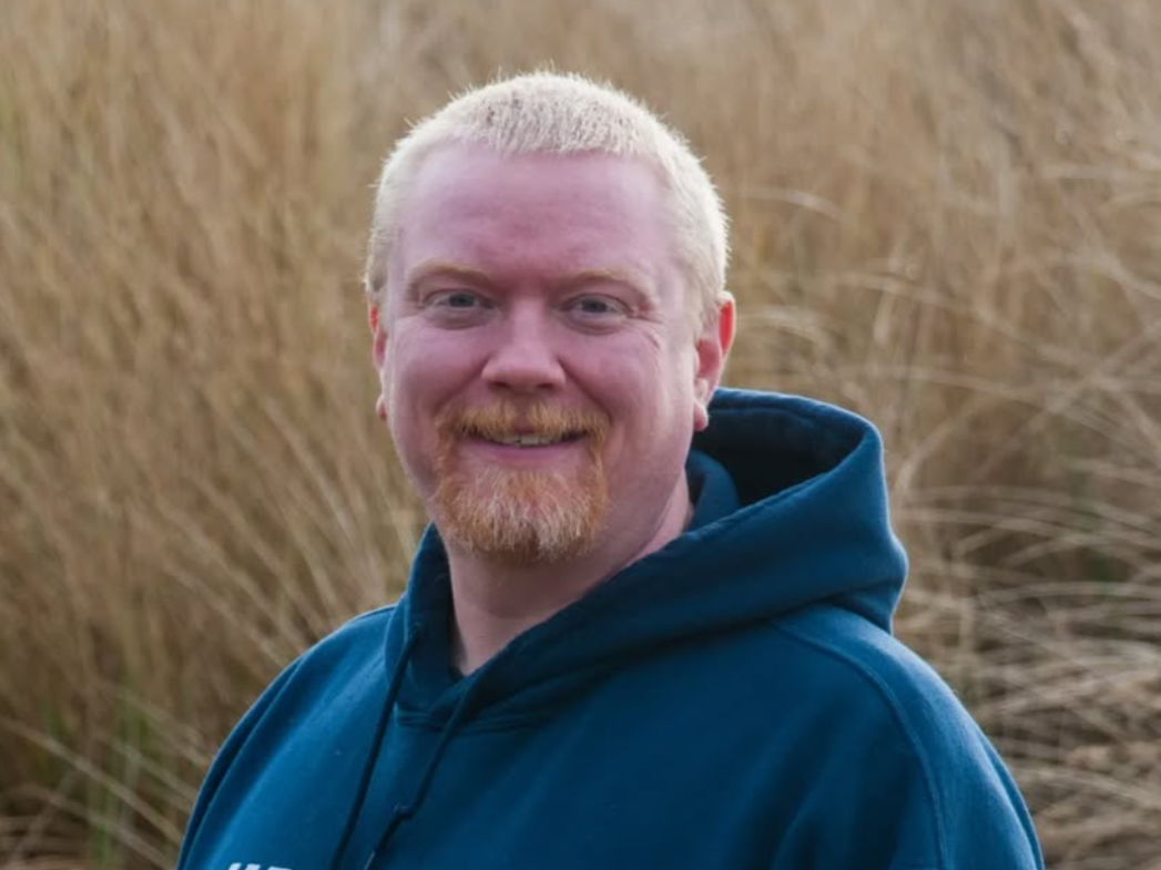 Jake Carlsen smiling outdoors in front of tall Oregon Coast dune grasses, wearing a blue hoodie.