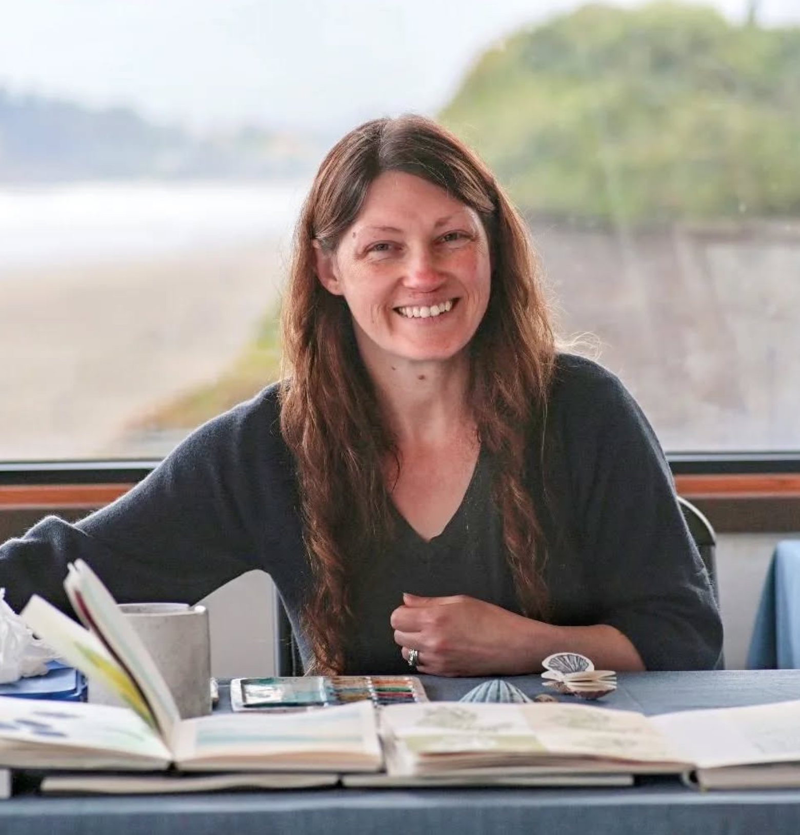 Christina Carlsen smiling while sitting at a table with books and papers, in front of a window with a scenic outdoor view of the Oregon Coast.
