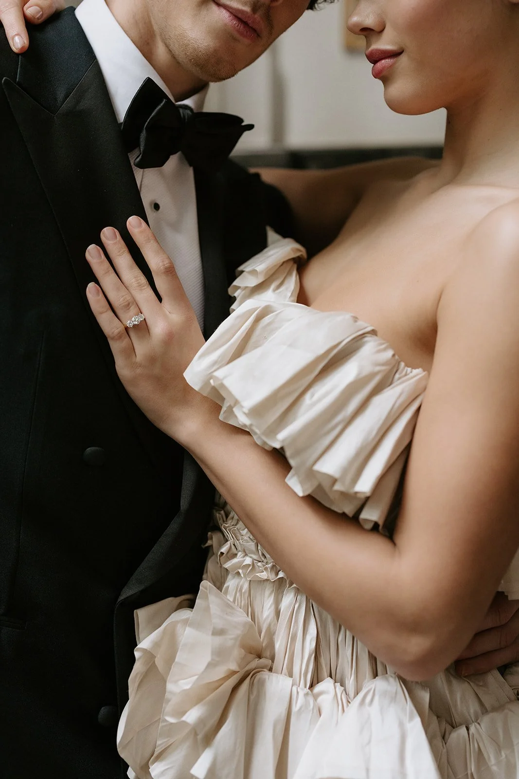 Close-up of a couple dressed in formal attire, with the woman wearing a cream-colored dress with ruffled sleeves, and the man in a tuxedo, showing their faces and upper bodies.