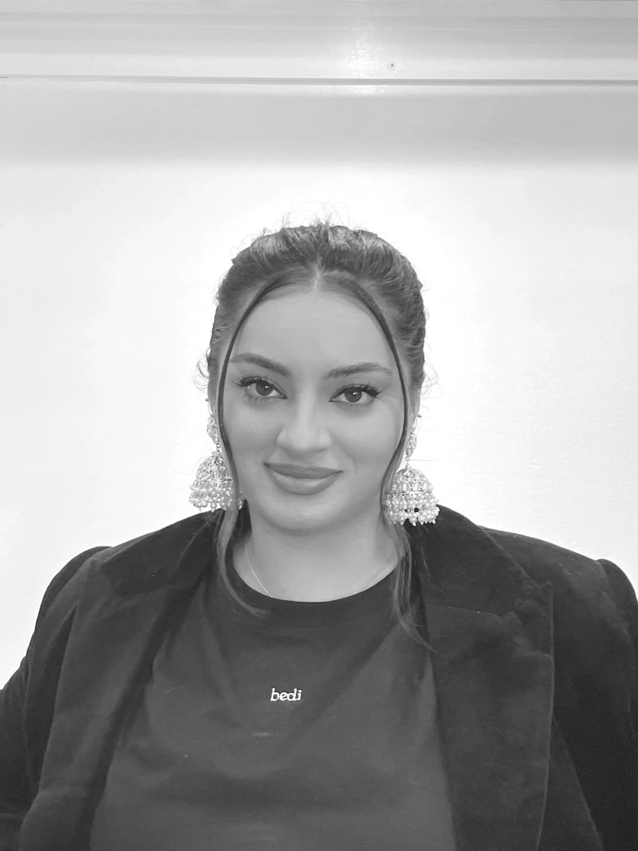 Black and white portrait of a person with long earrings, wearing a black jacket and a shirt with "bedi" written on it, neutral background.