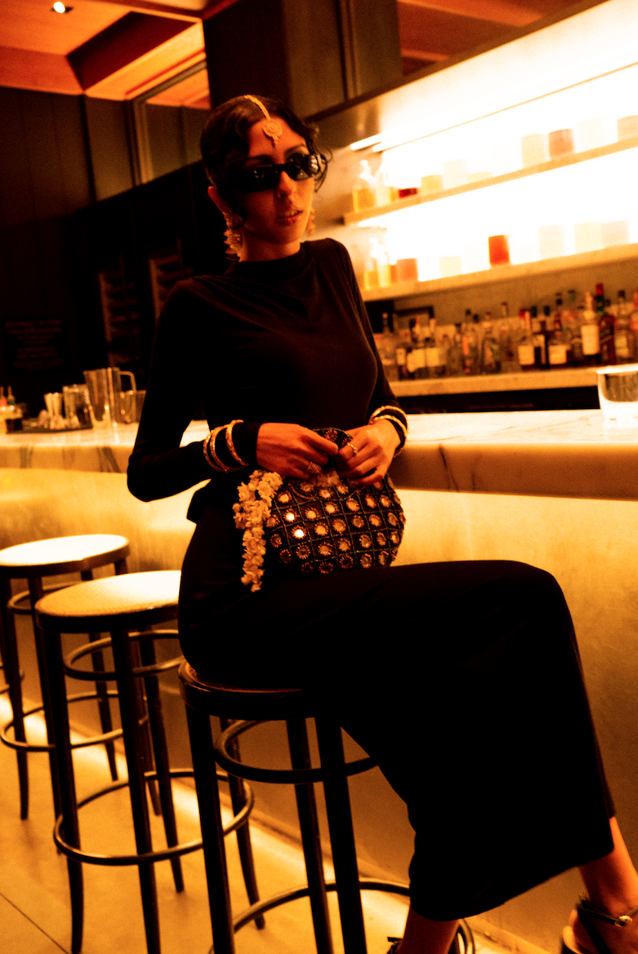 Stylish person with sunglasses sitting at a bar counter, holding a decorative handbag, in a dimly lit bar setting with bottles on shelves.