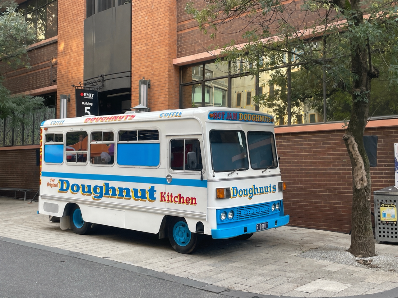 A mobile food truck designed to look like a vintage doughnut shop, parked on a sidewalk in front of a brick building with large windows. The truck has colorful signs advertising doughnuts, coffee, and hot jam doughnuts, with bright blue and white colors.