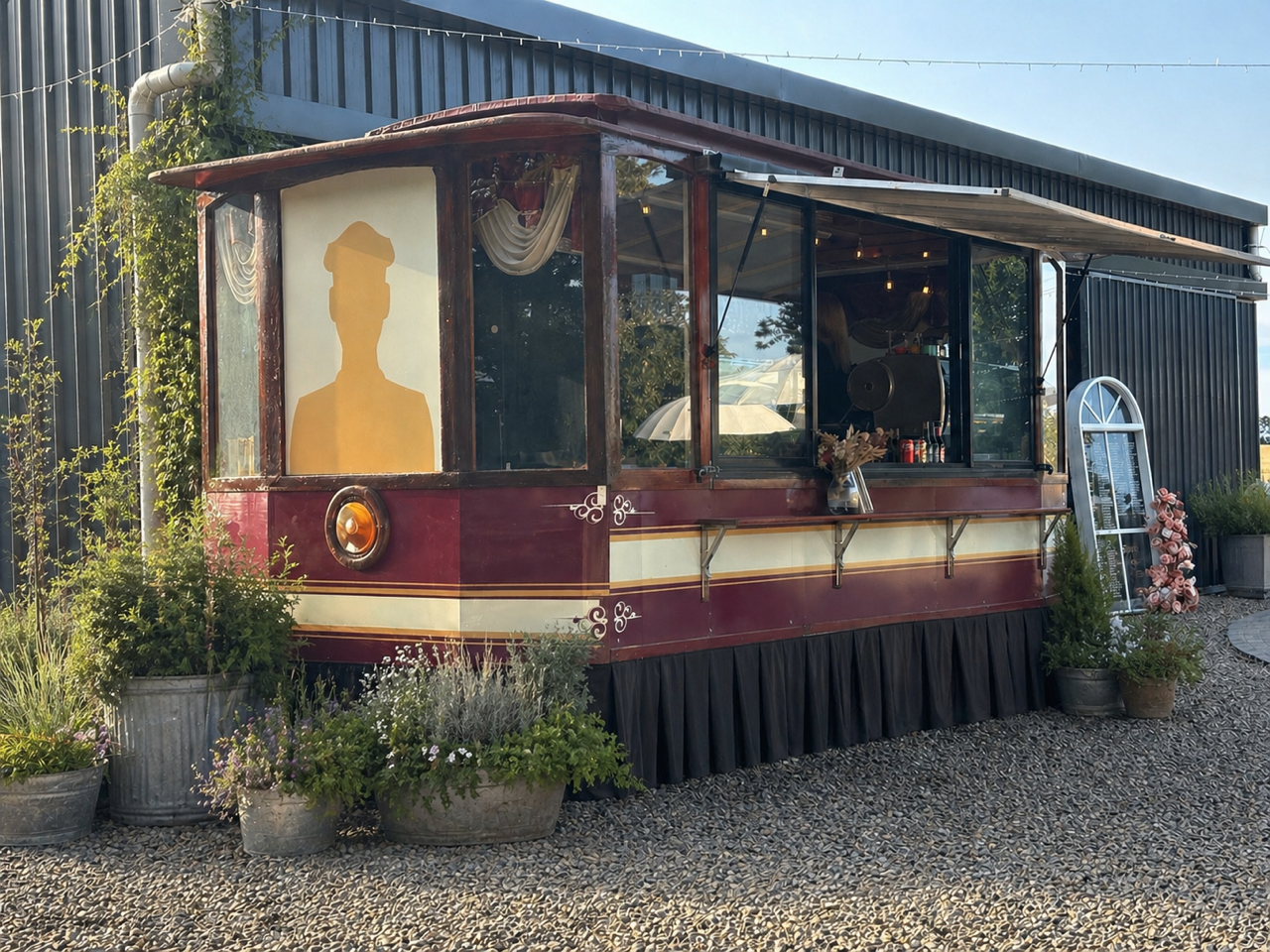 Vintage food truck with a silhouette of a person in hat on the window, surrounded by potted plants and flowers outside, with a modern building in the background.