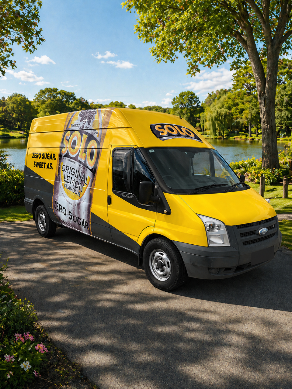 A yellow delivery van parked on a paved pathway in a park with trees and a pond, advertising Solo lemon-flavored drink with zero sugar.