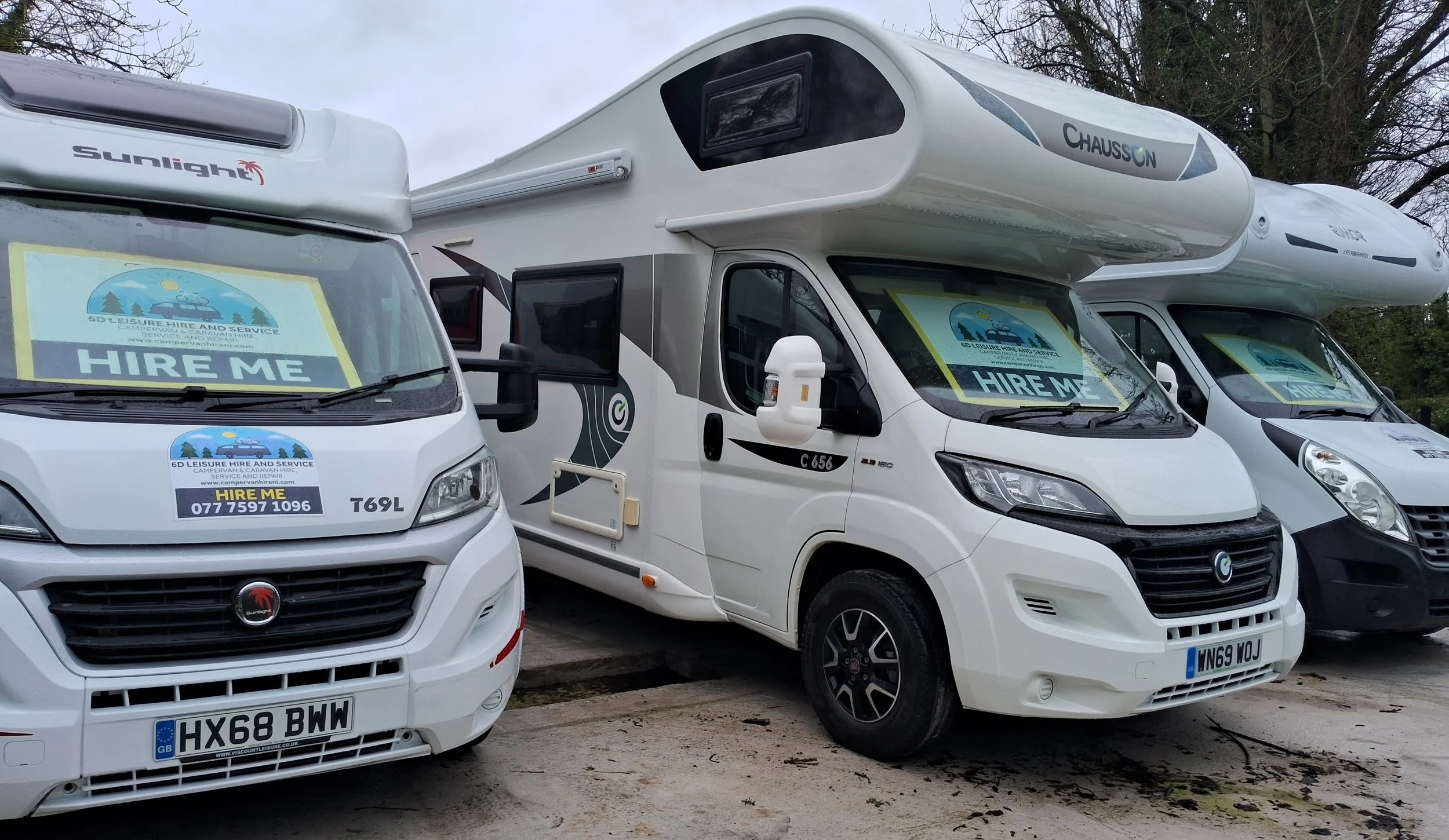 Three camper vans parked side by side outdoors. Each van has a sign in the windshield that says 'HIRE ME' and contact information for a leisure hire and service company.