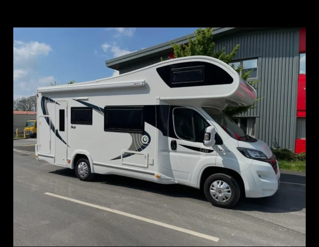 White recreational vehicle (RV) parked on the side of a road near a building with gray metal walls and red accents.