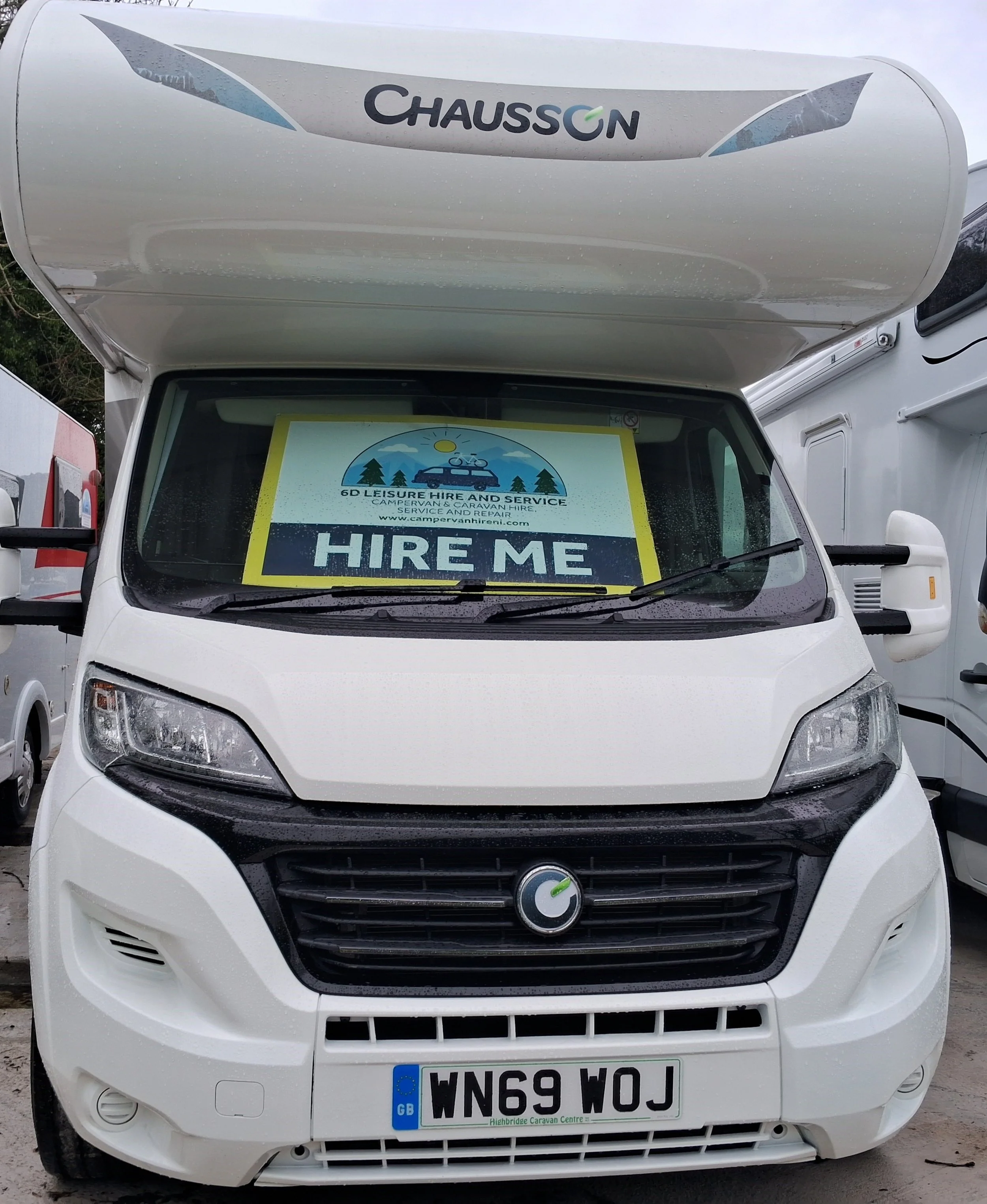 Front view of a white motorhome with a sign in the front window stating 'Hire Me' by 6D Leisure Hire and Service. The vehicle has a 'Chausson' logo on the top front.