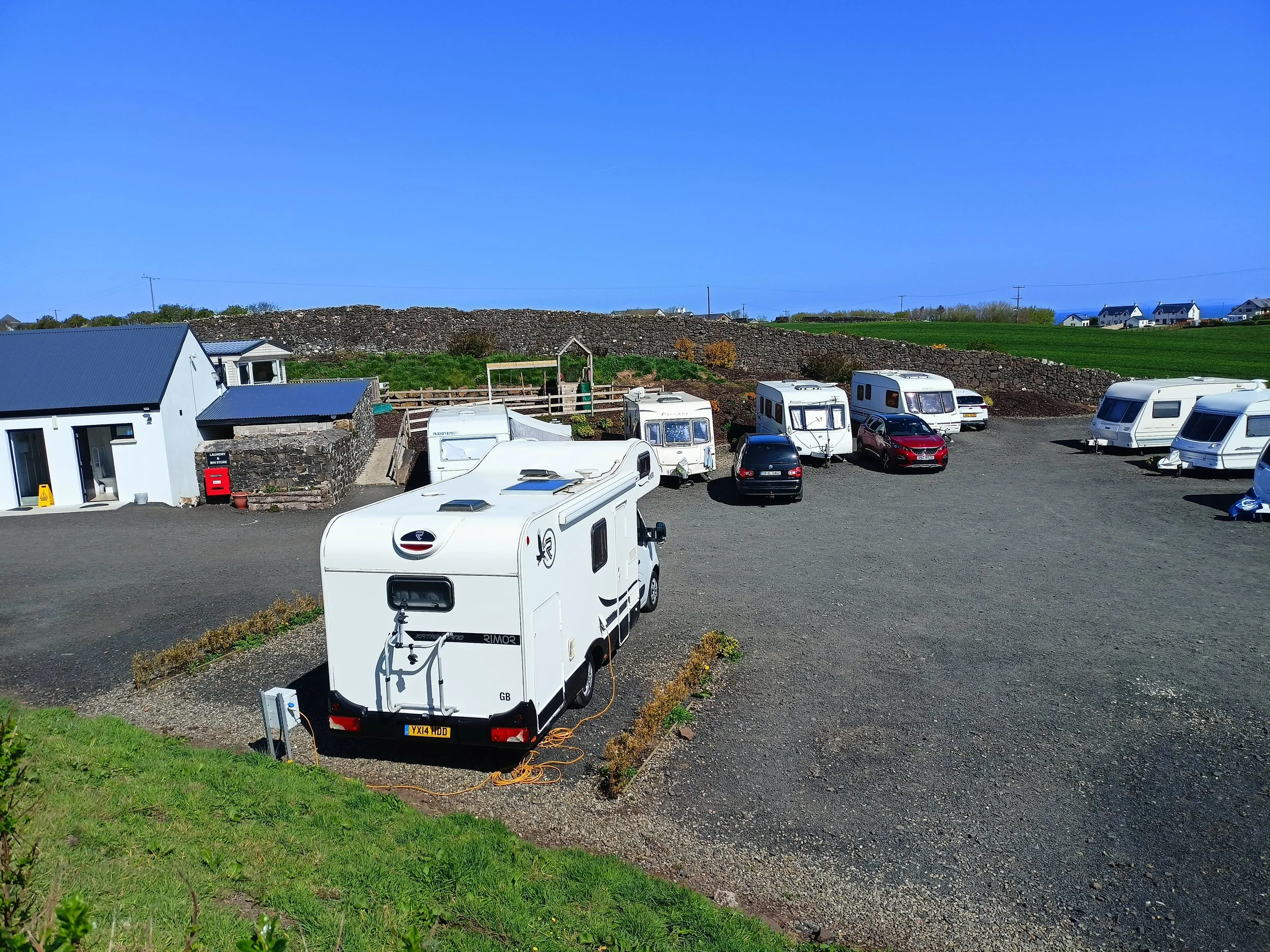 A parking lot with several white camper vans and a few cars under a clear blue sky, surrounded by green fields and houses in the background.