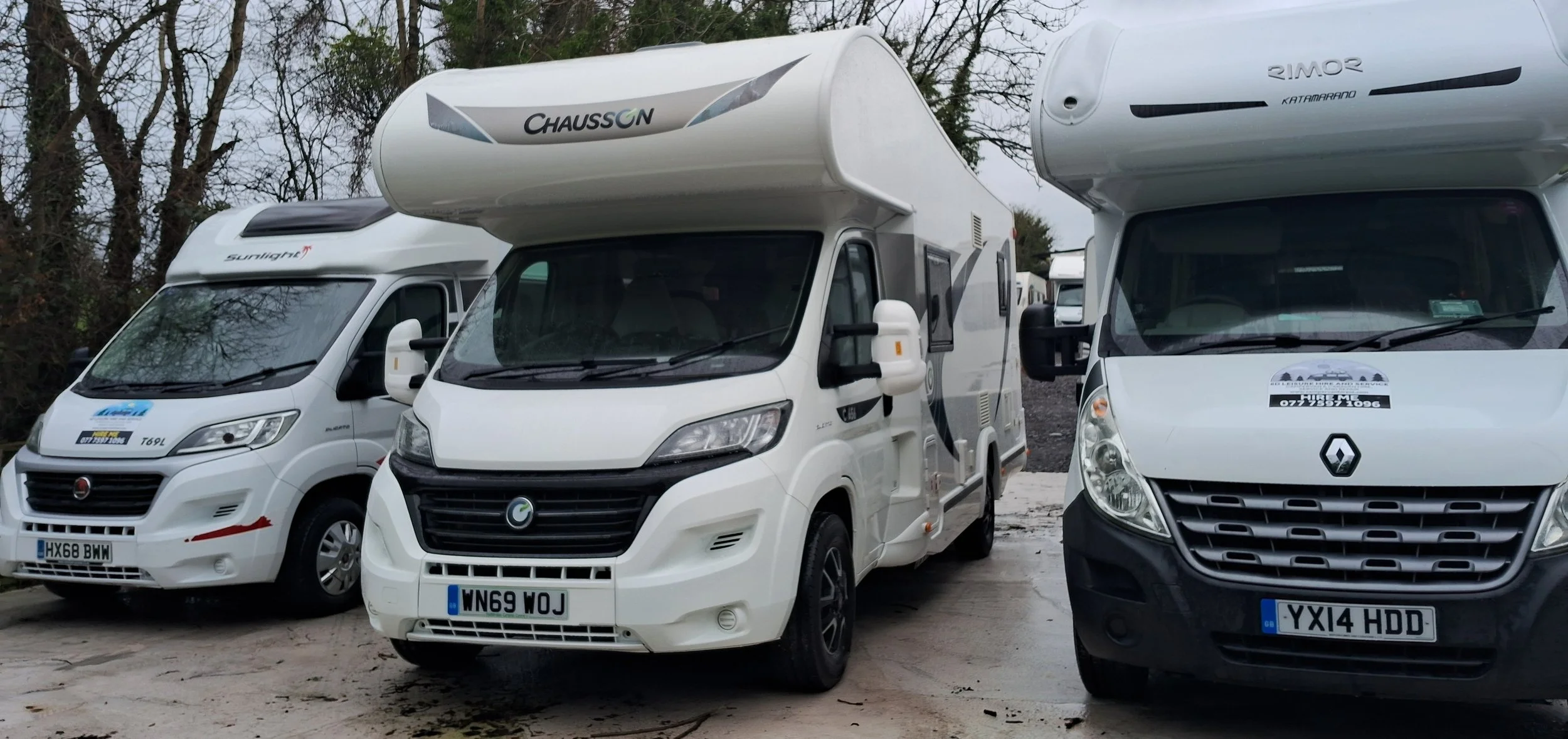 Three white camper vans parked outdoors on a cloudy day, with trees in the background.