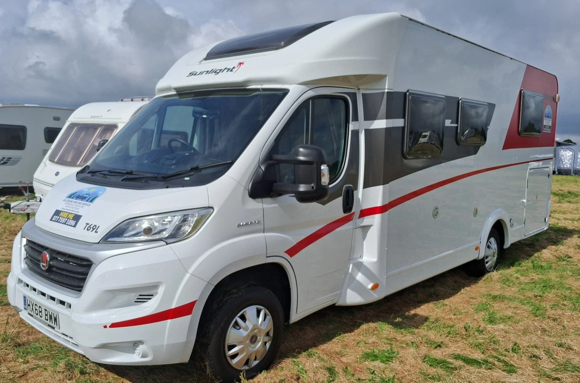 A white vehicle motorhome with black and red stripes on a grassy field with other RVs in the background, under a partly cloudy sky.