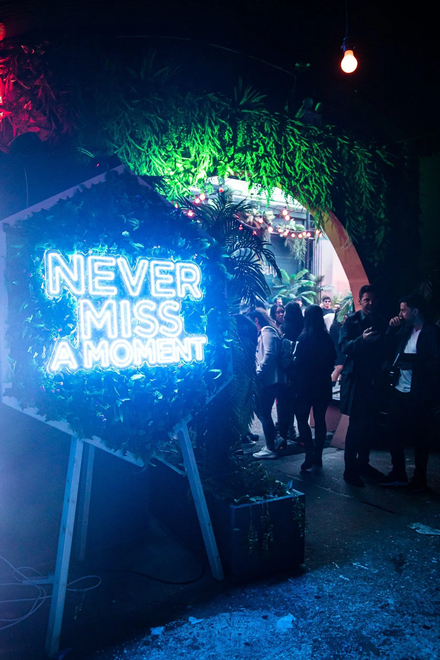 A neon sign that reads 'Never Miss A Moment' in bright blue, with people standing around and socializing under a canopy of green foliage and red string lights at night.