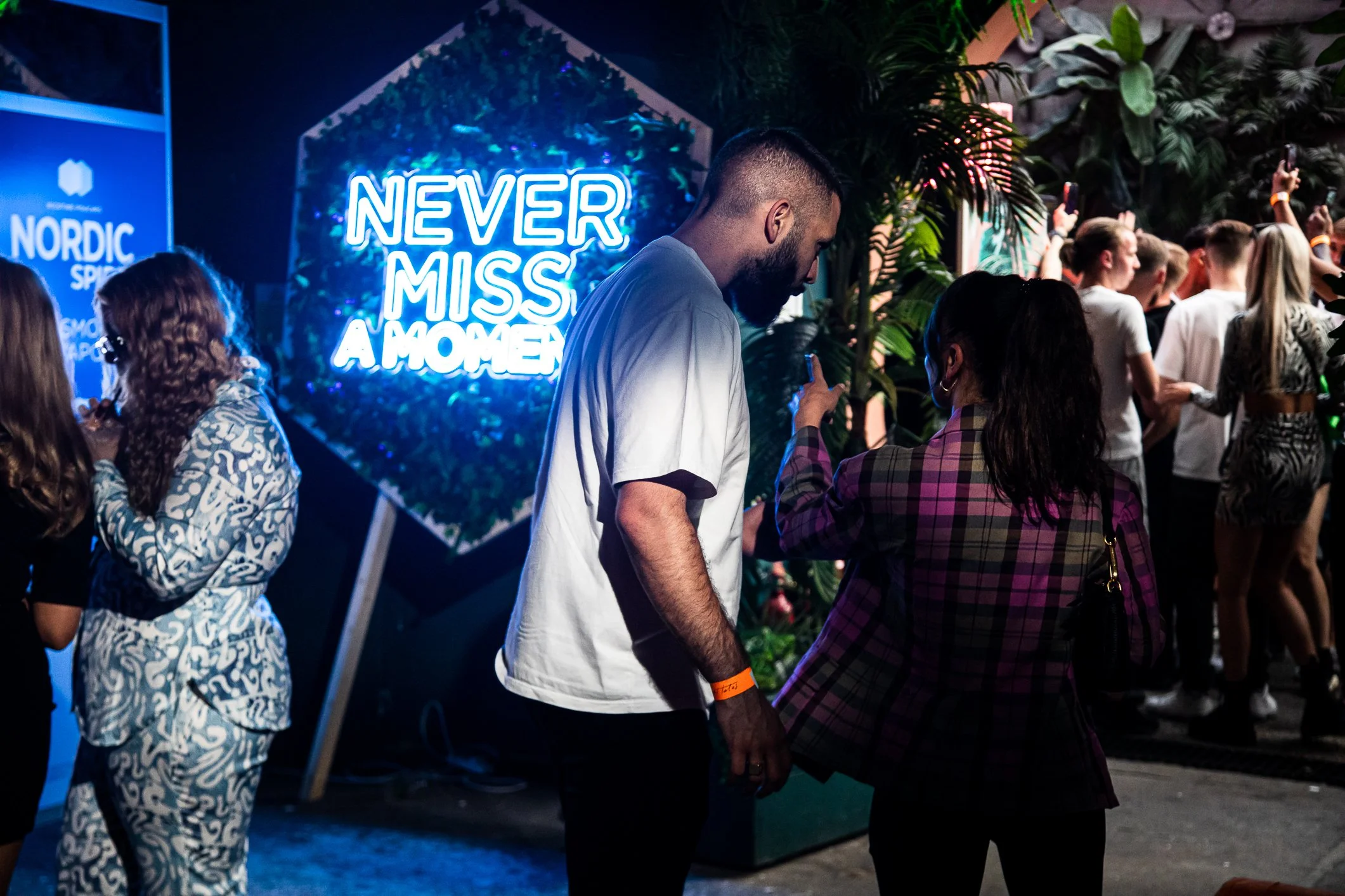 Two people dancing at an event with a neon sign that reads 'Never Miss a Moment', surrounded by other party-goers and lush greenery in the background.