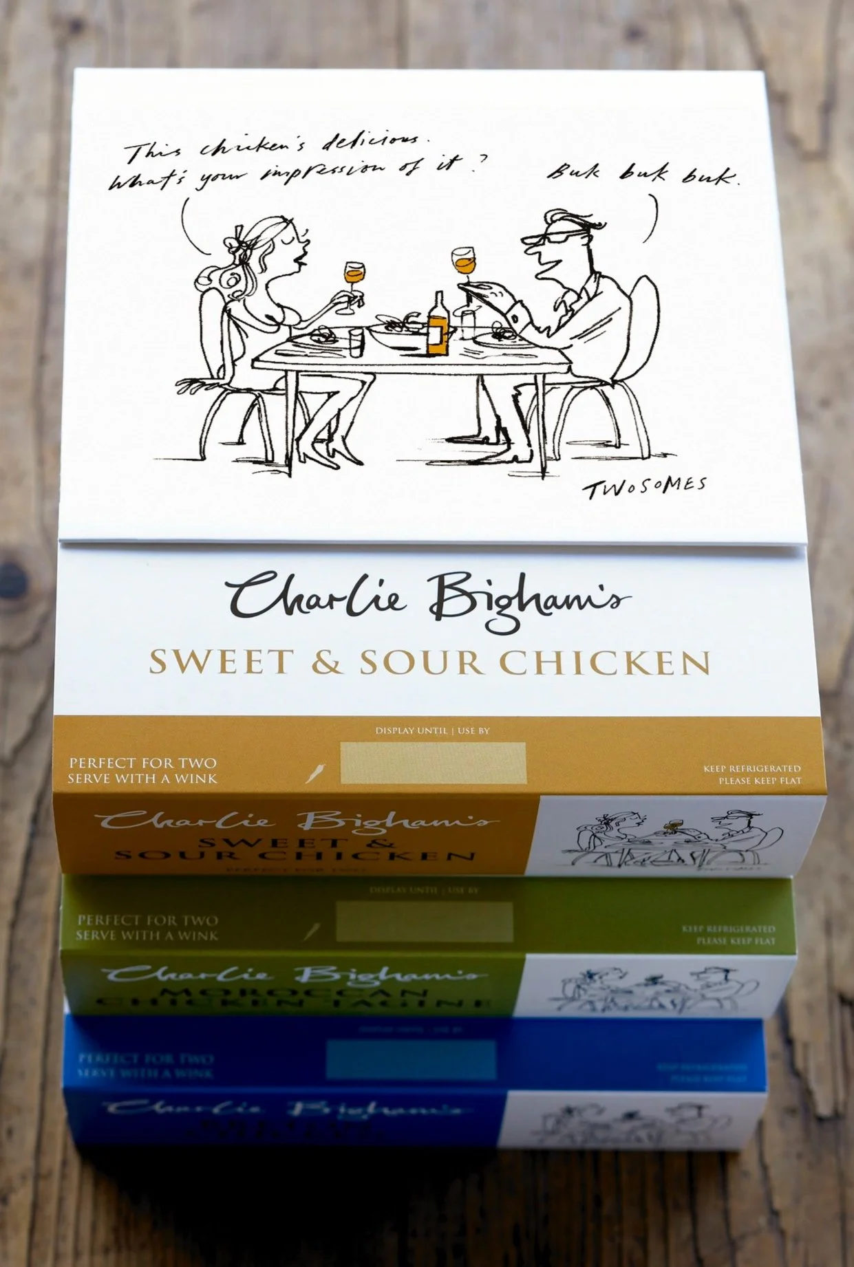 Boxes of Charlie Bigham's Sweet & Sour Chicken meal in three colors: orange, green, and blue, stacked on a wooden surface with illustration of a couple having dinner on the top box.