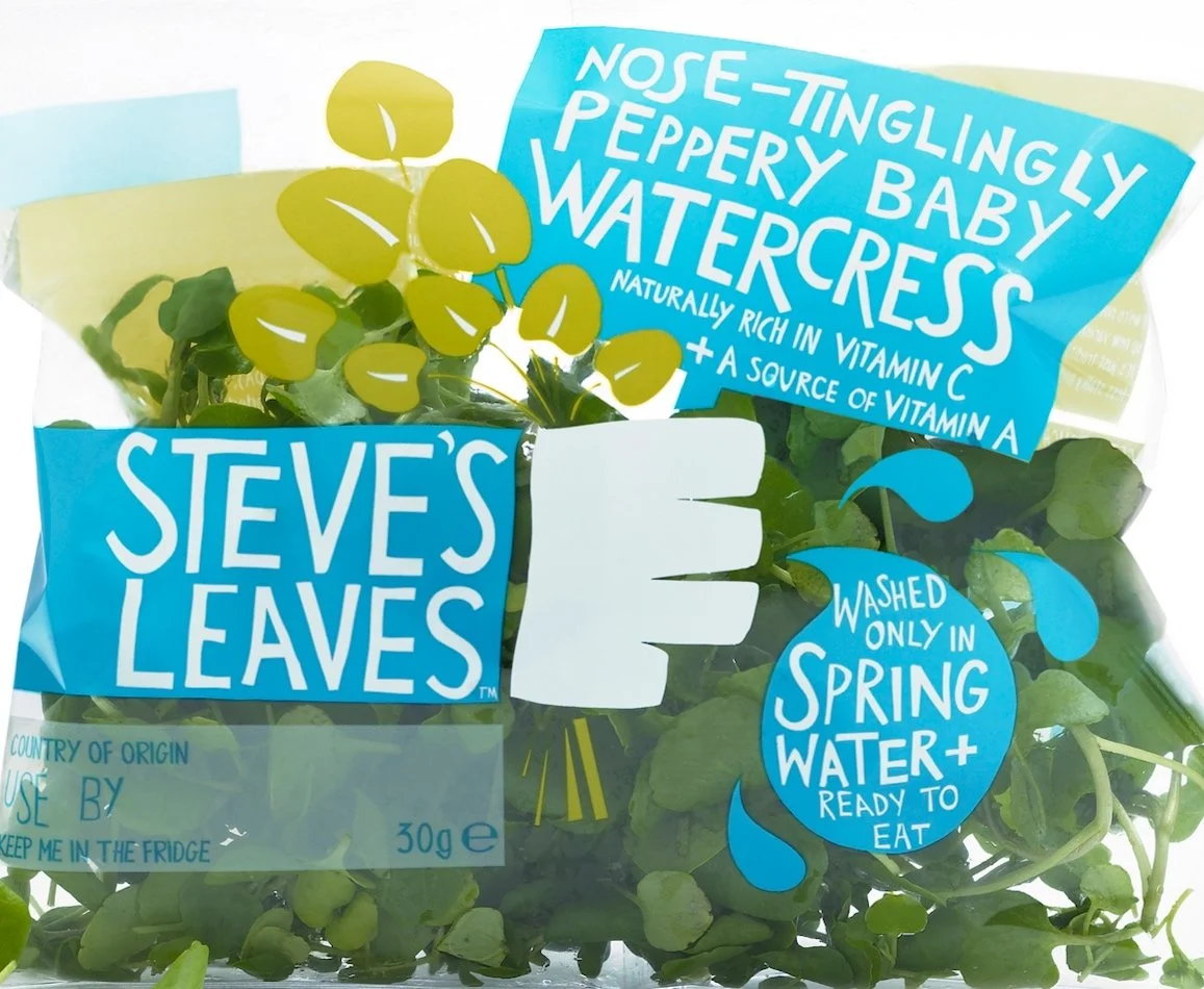 Fresh watercress in clear plastic bag with blue labels indicating it's grown under spring water and contains vitamins, branded as Steve's Leaves.