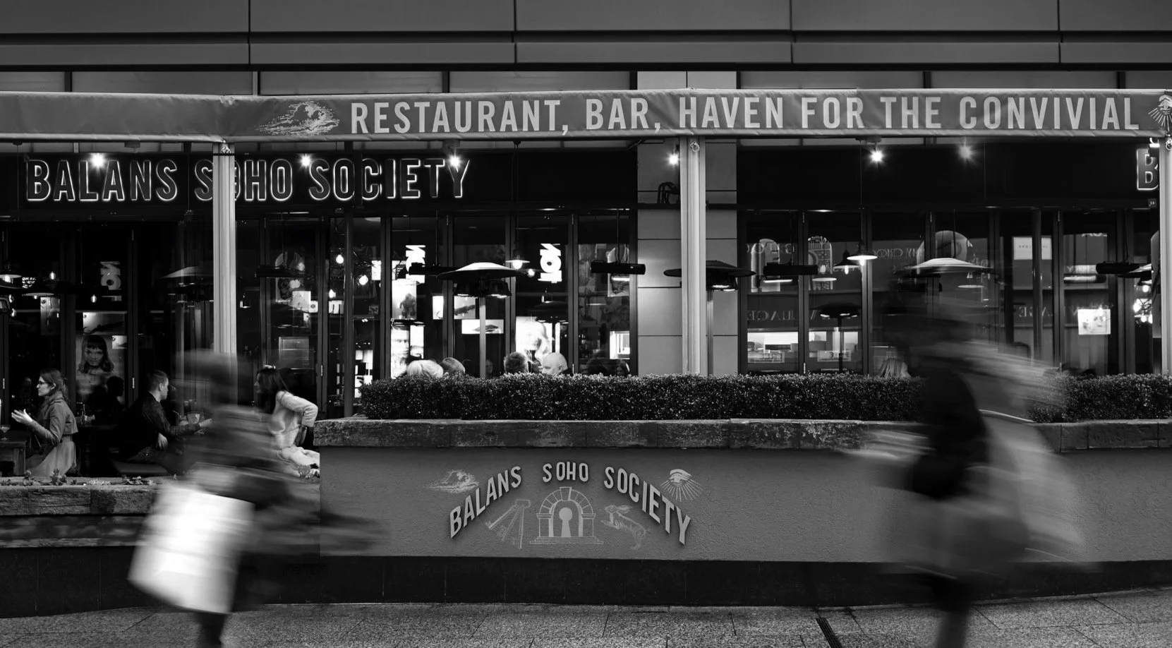 Night scene of Balans Soho Society restaurant and bar with people sitting outside and pedestrians riding bikes in front, black and white photograph.