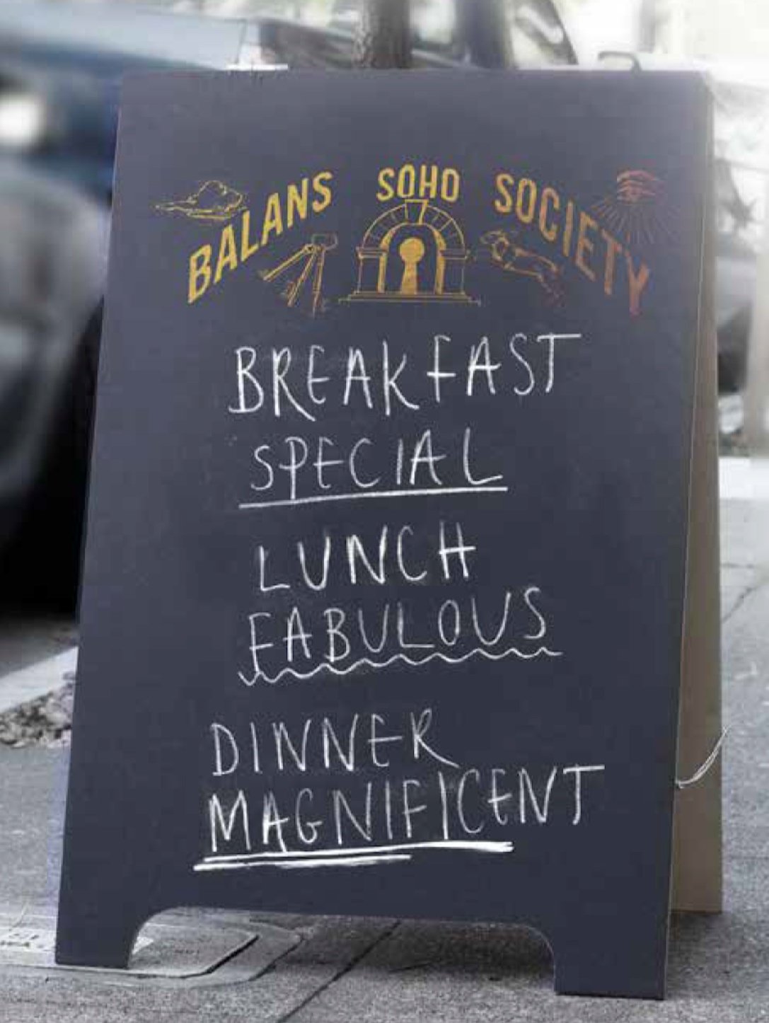 Black chalkboard sign with yellow and white text, advertising a breakfast, lunch, and dinner special at Balans Soho Society.