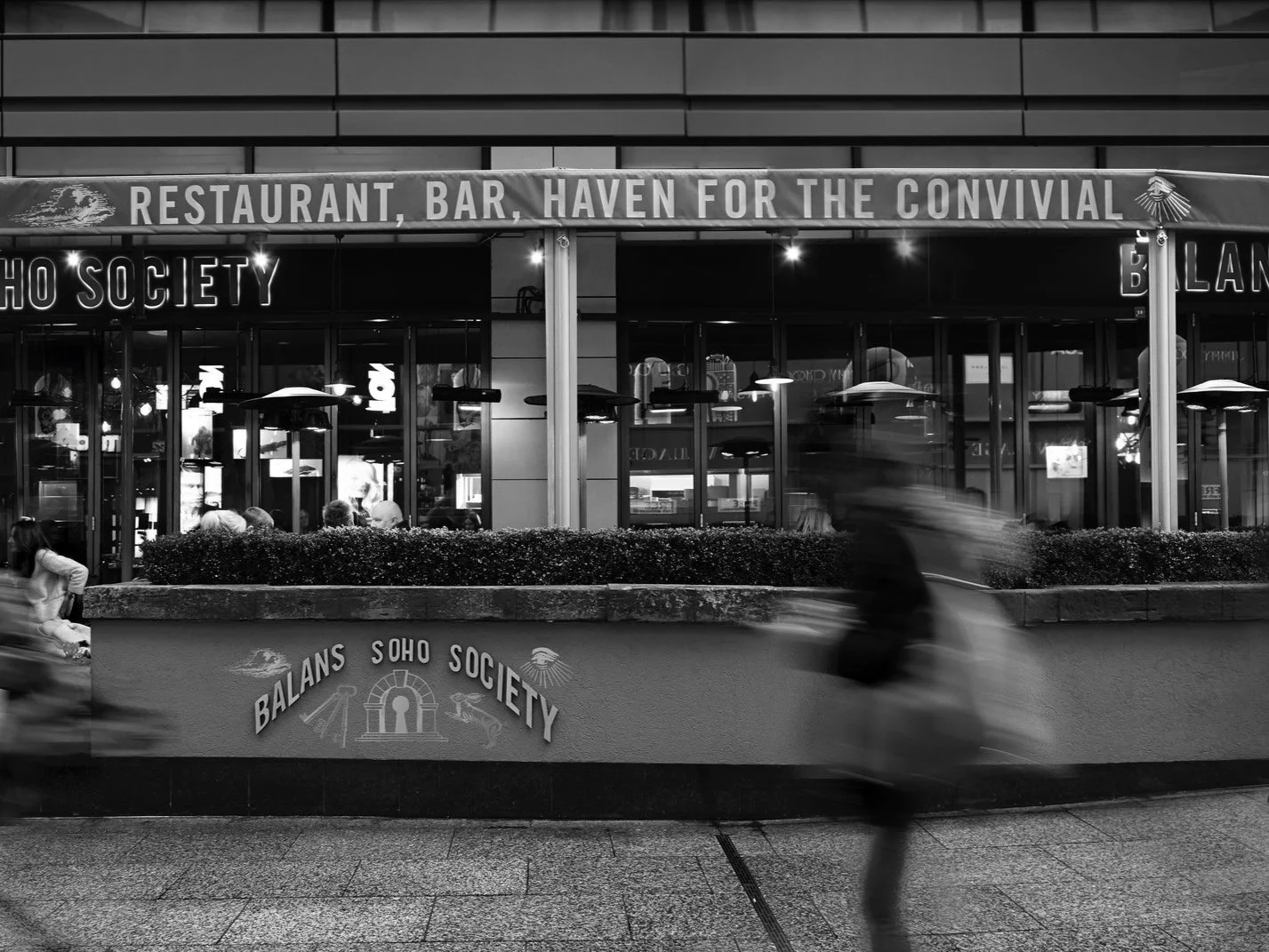 Black and white photo of a Soho Society restaurant and bar with outdoor heaters and people inside, with a person riding a scooter blurred in motion in front.