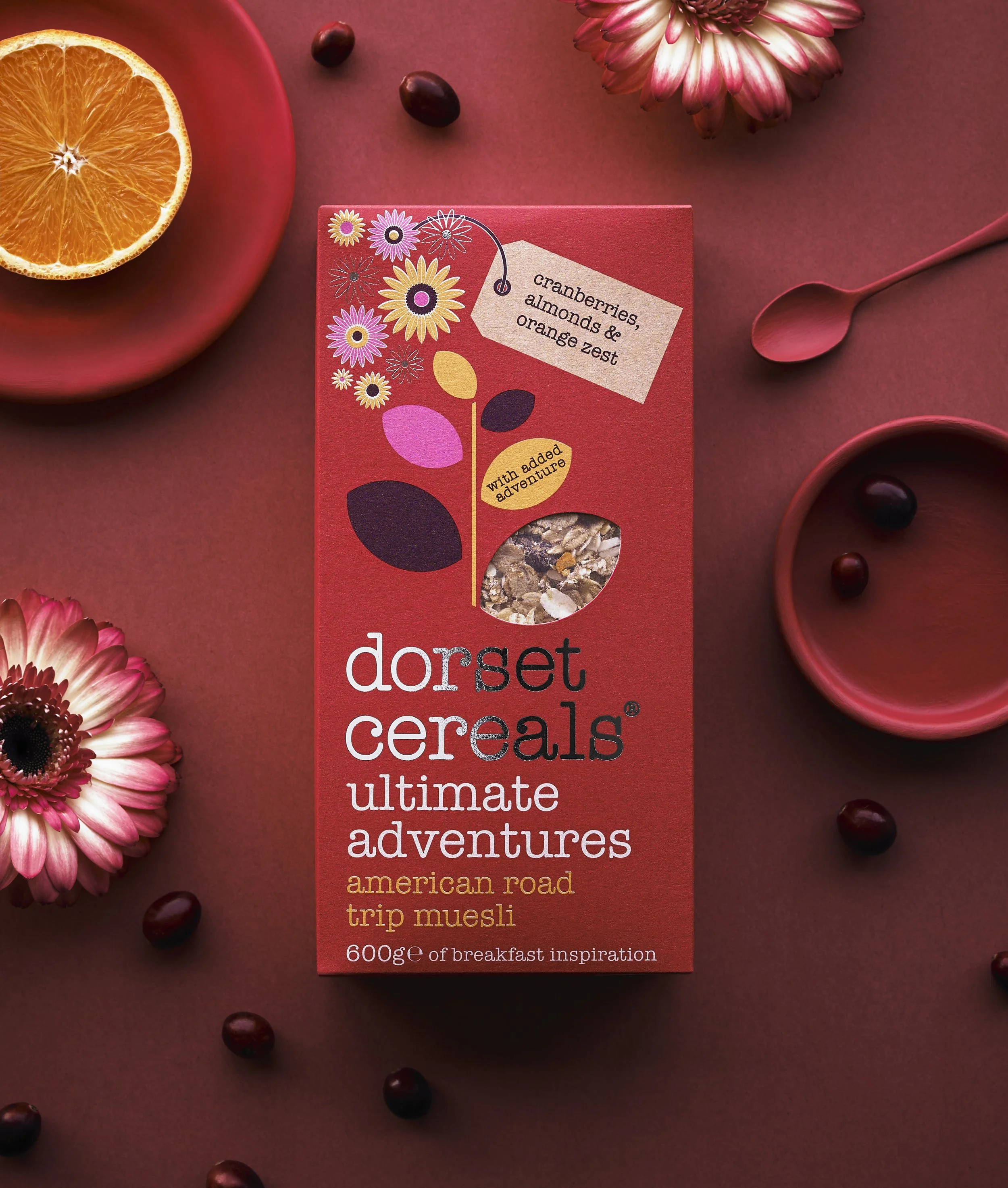 A red box of Dorset Cereals Ultimate Adventures American Road Trip Muesli surrounded by pink and red flowers, a halved orange, a small bowl, and scattered berries on a red background.