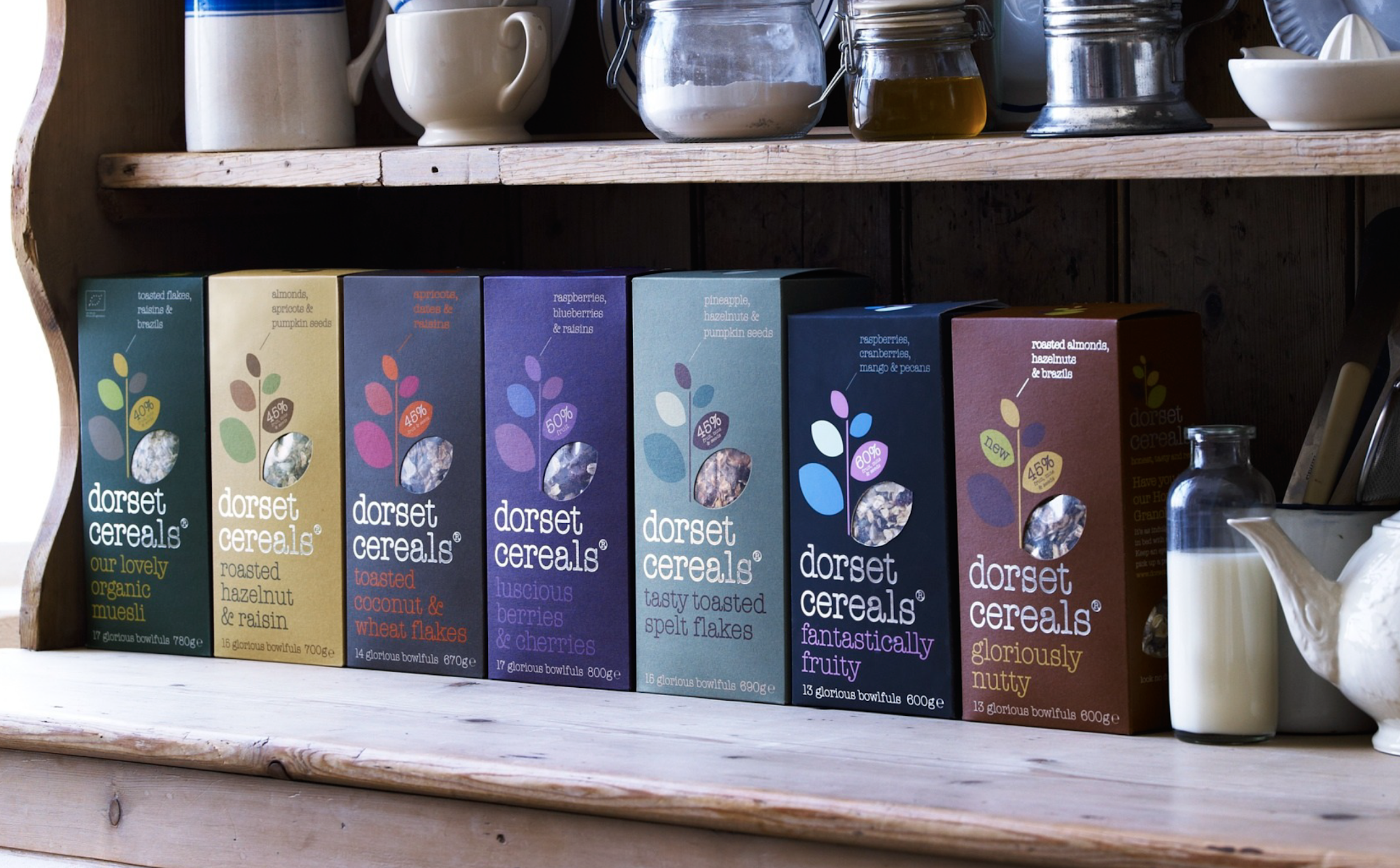 Shelf of various Dorset Cereals boxes with different flavors, including organic muesli, hazelnut and raisin, toasted coconut and wheat flakes, berries and cherries, toasted spelt flakes, fruity, and Nutty, on a wooden shelf.
