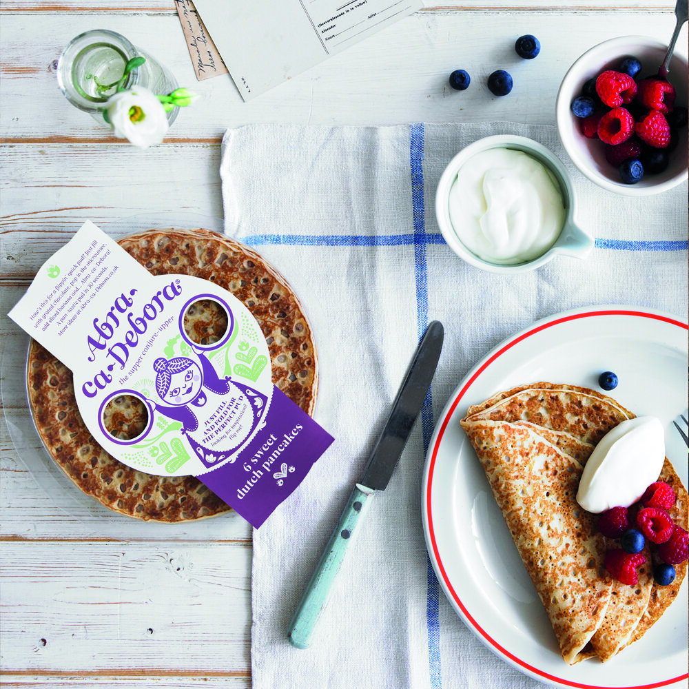 A breakfast setting with a plate of crepes topped with whipped cream and mixed berries, a bowl of additional berries, a cup of yogurt, a small vase with a white flower, and a package of Dutch pancakes on a white wooden table.