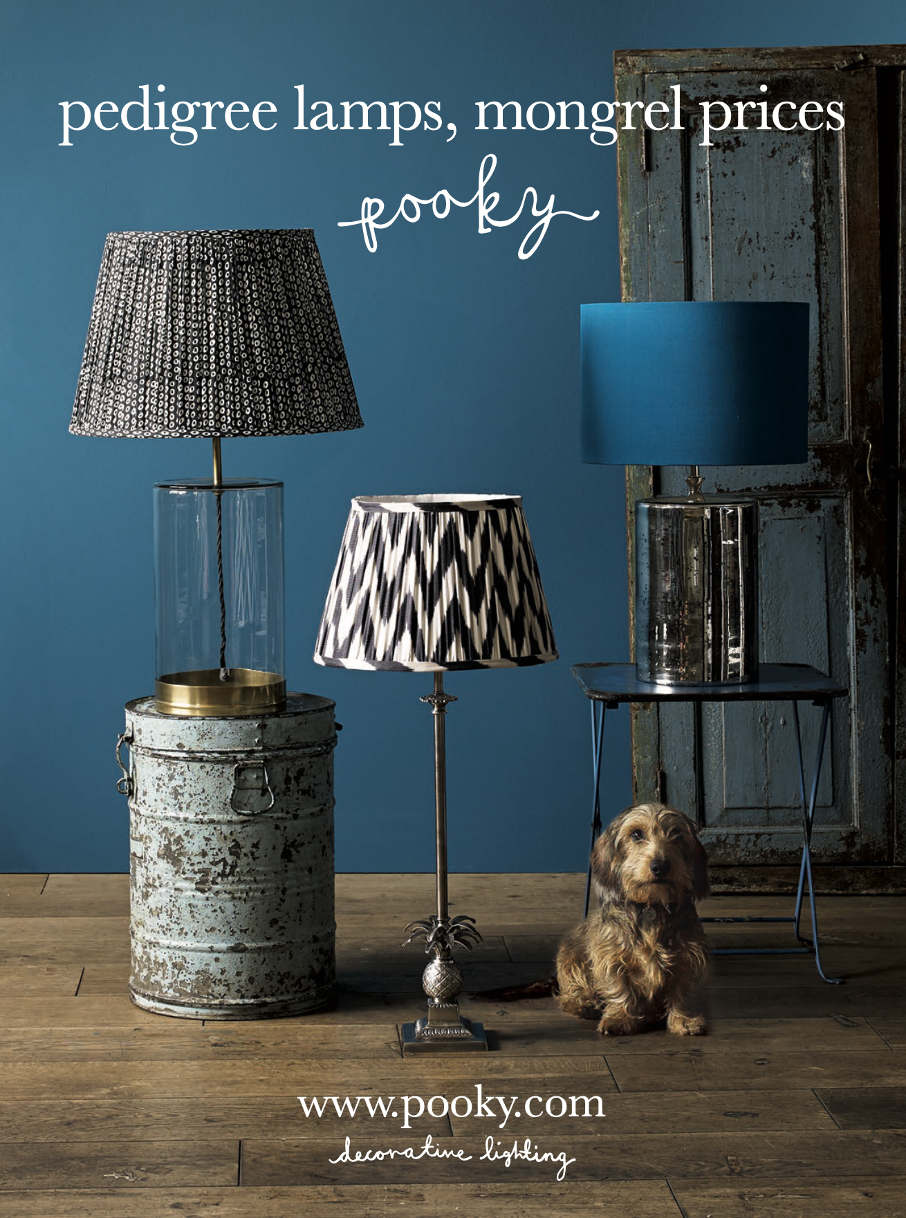 Arrangement of three pedestal lamps with patterned shades, one on a vintage metal stand, another in a glass base, and the third on a small table, with a small dog sitting on a wooden floor against a blue wall, advertising Pooky decorative lighting.