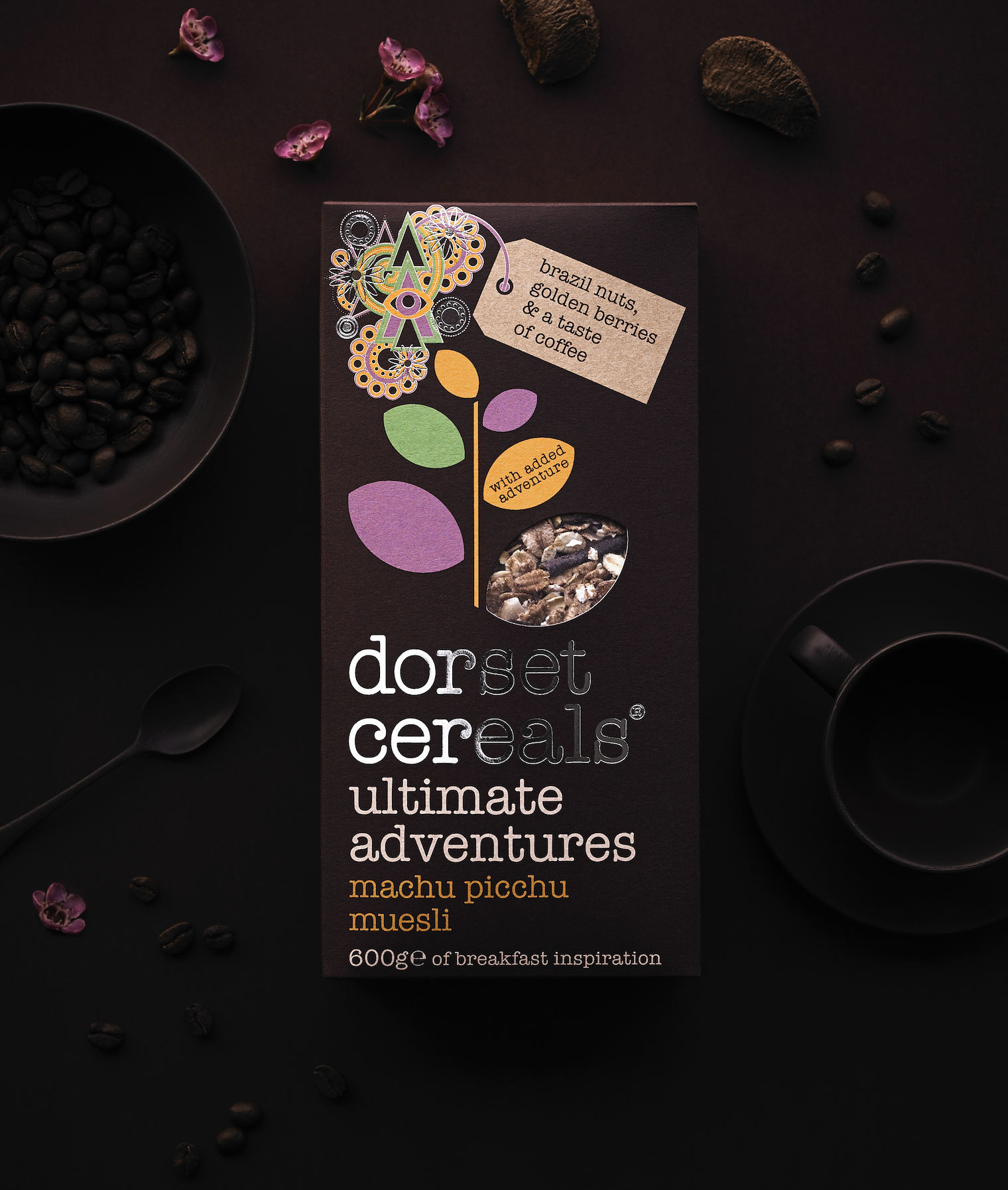 Package of Dorset Cereals Ultimate Adventures Machu Picchu Muesli surrounded by coffee beans, a cup of coffee, a spoon, and purple flowers on a dark background.