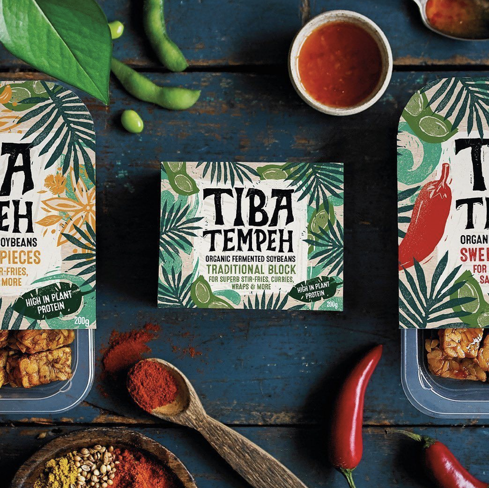 Center package of Tiba Tempeh, surrounded by chili peppers, a wooden spoon with red spice, a small bowl of red sauce, and other food items on a rustic blue wooden surface.