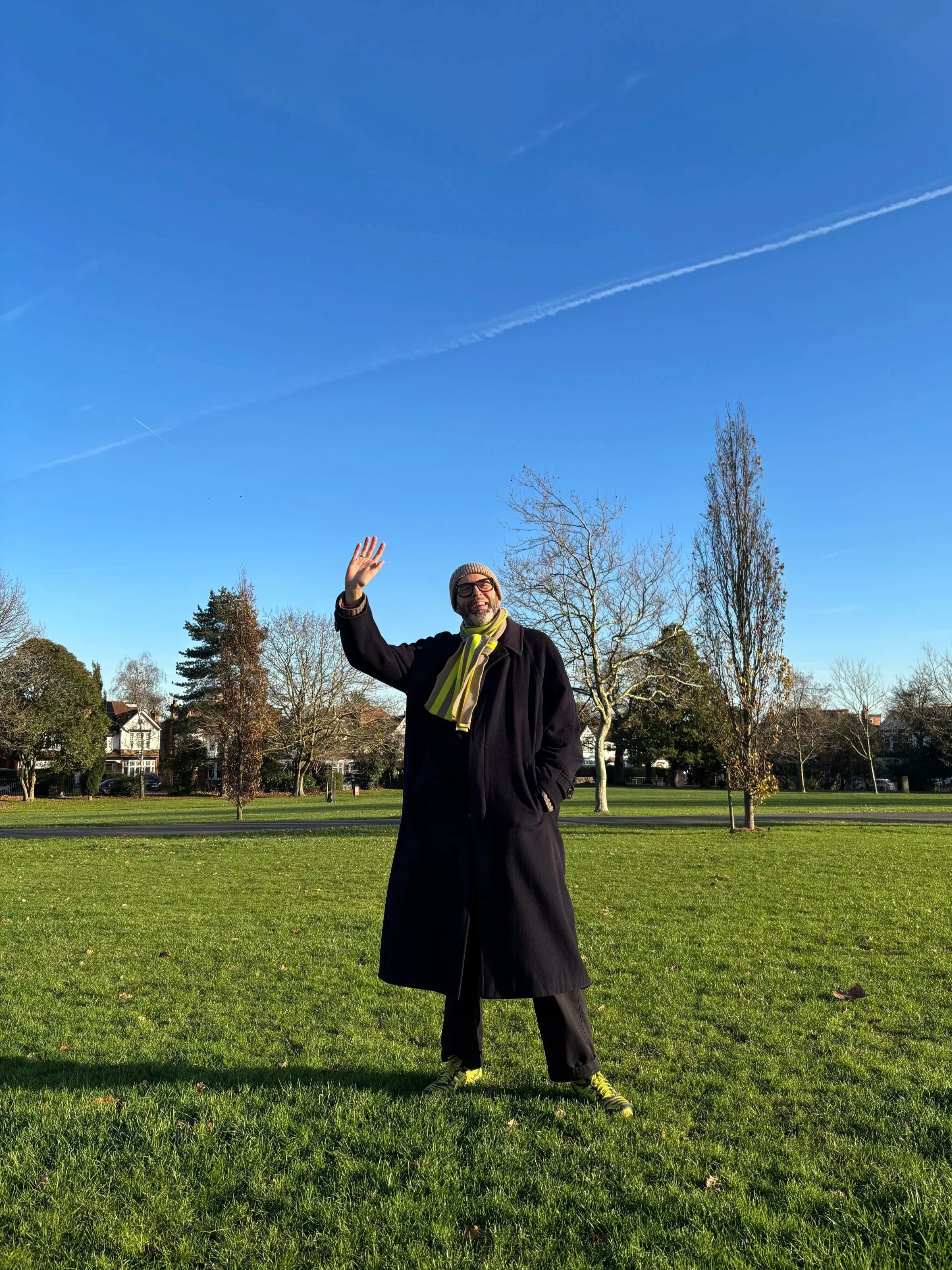 A man with glasses, a gray beard, and a gray beanie wearing a black coat and a striped scarf, waving and smiling in a park with green grass, leafless trees, and houses in the background under a clear blue sky with contrails.
