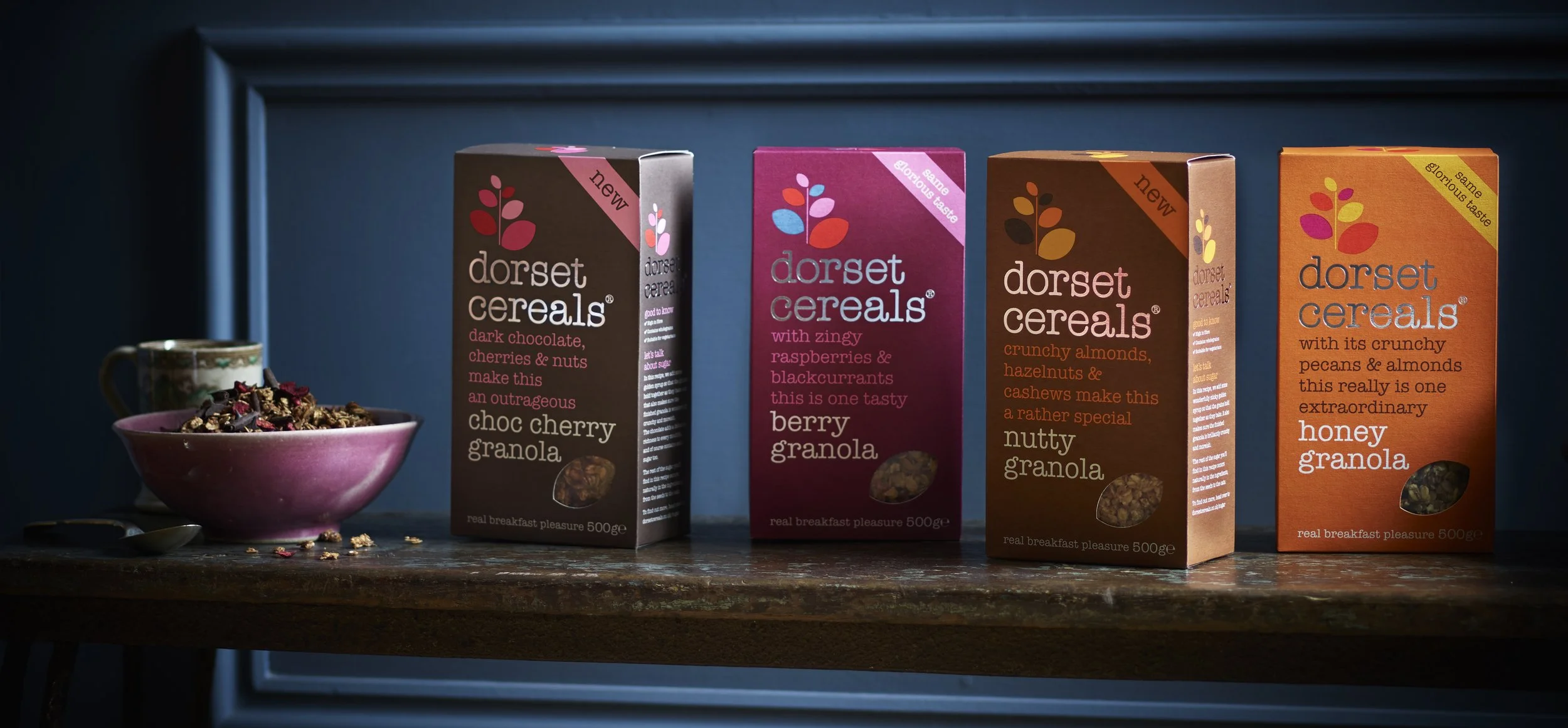 Four boxes of Dorset Cereals granola on a wooden surface, with a pink bowl of granola and scattered granola pieces nearby.