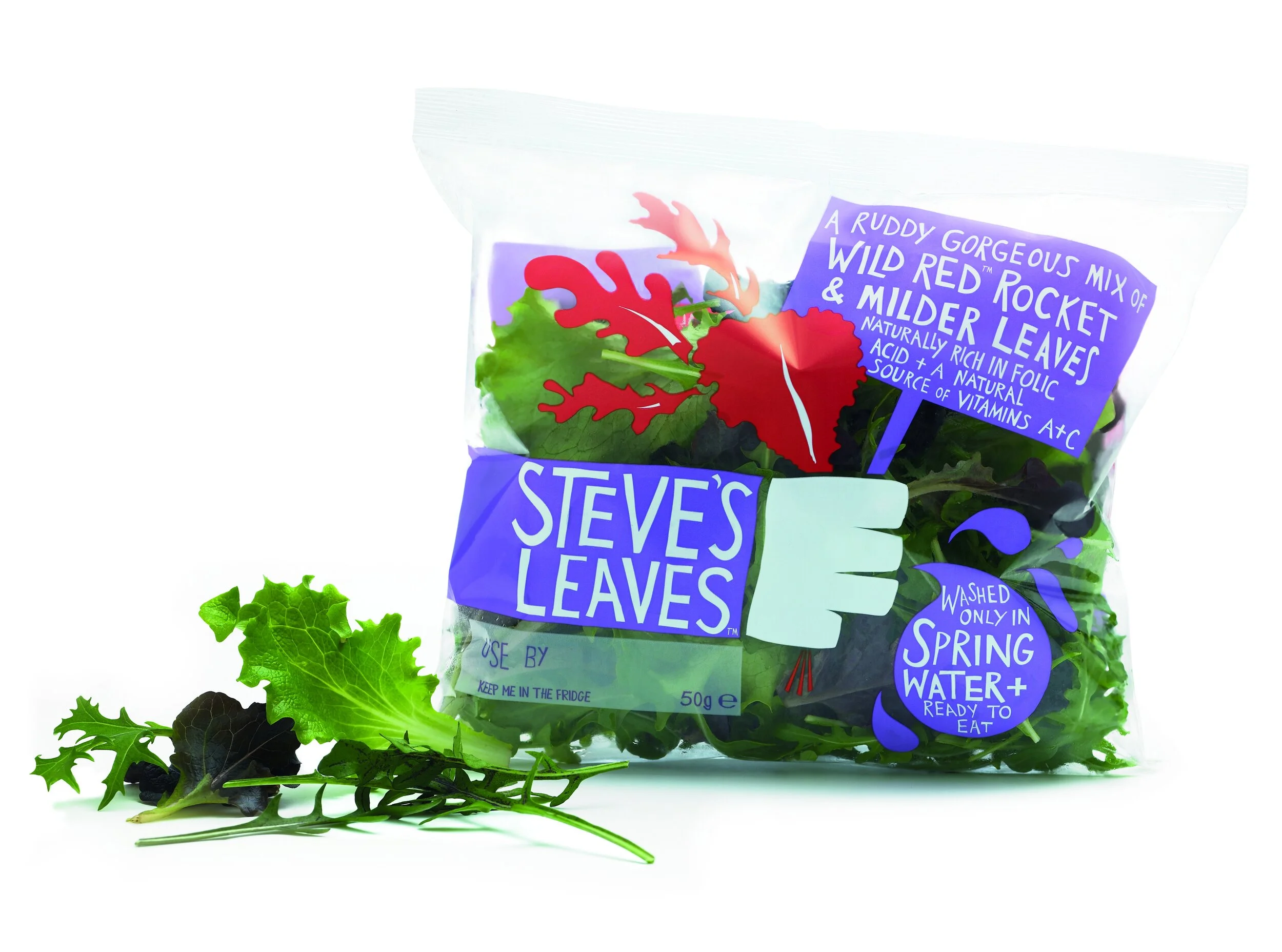 A clear plastic bag of mixed salad greens with a label reading "Steve's Leaves," and a purple sign indicating it contains a mix of wild red rocket and milder leaves, washed only in spring water and ready to eat, with some loose leaves outside the bag