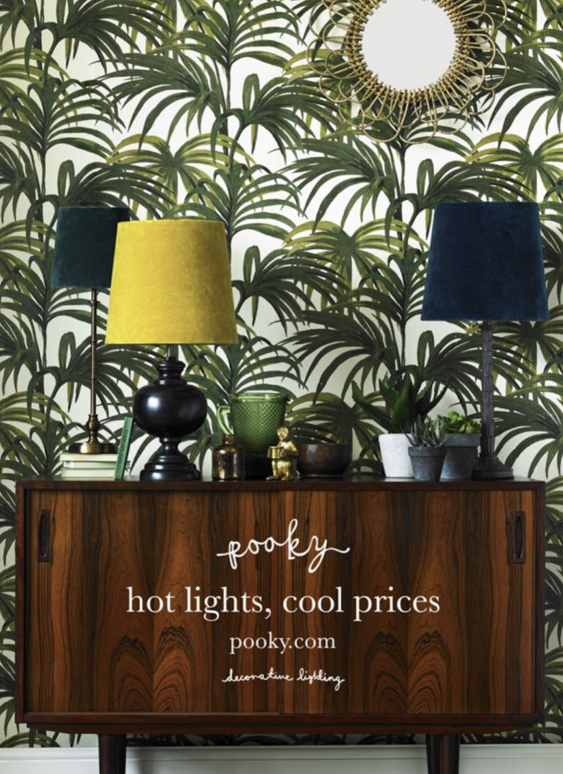 A press ad for Pooky lighting. A wooden sideboard with decorative items including two green lampshades, small plants, and decorative objects, set against a wall with green palm leaf wallpaper and a round mirror with a gold frame. Copy by Lee Anderson