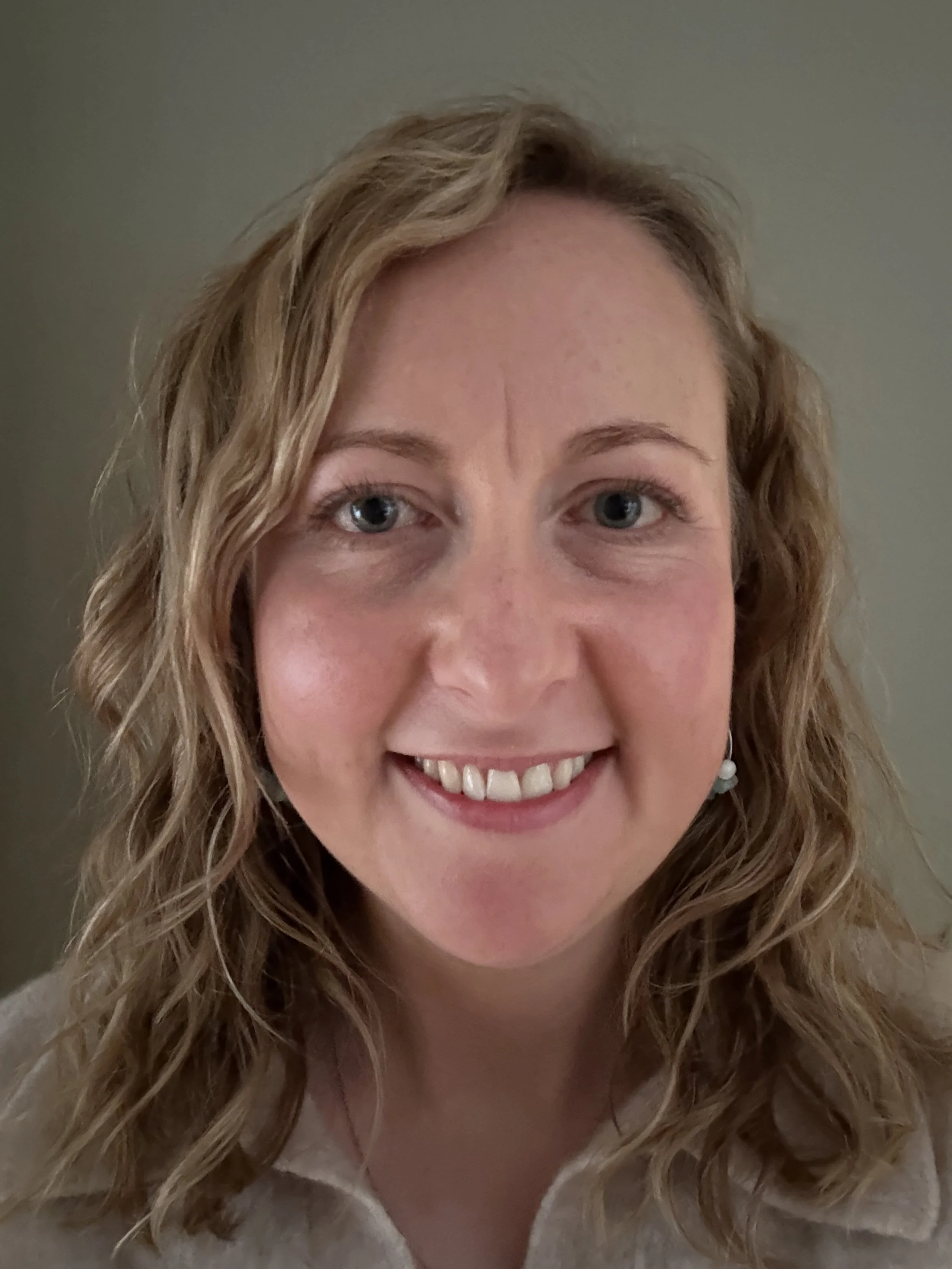 Close-up of a smiling woman with wavy blonde hair and blue eyes, wearing a white top and pearl earrings, with a plain background.