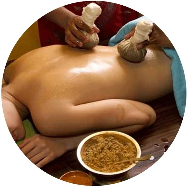 A person receiving a traditional massage, lying face down with their back exposed. Two individuals are applying massage sticks to the person's back, and a bowl of herbal massage paste is placed nearby.