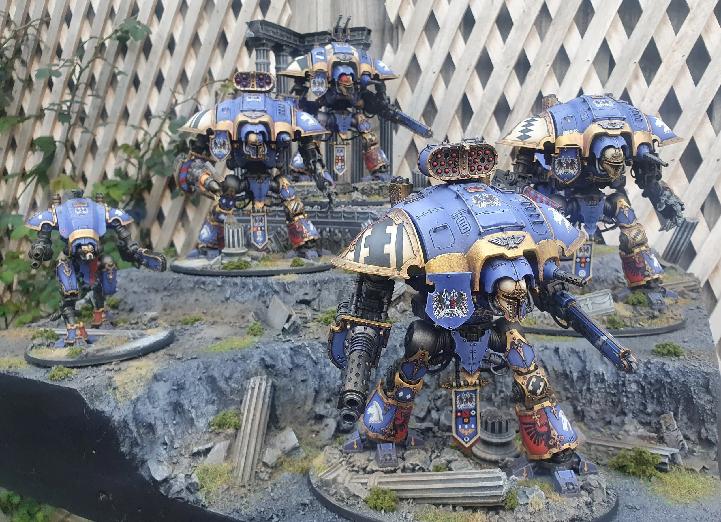 Miniature models of futuristic armored robots in blue and gold colors on a rocky terrain with plant elements, wooden fence in background.