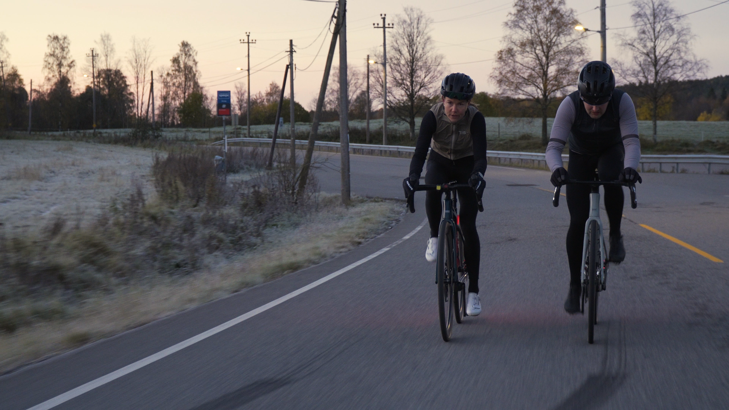Two cyclists wearing helmets and sporty clothing riding on a winding road during dawn or dusk, with trees and utility poles in the background.