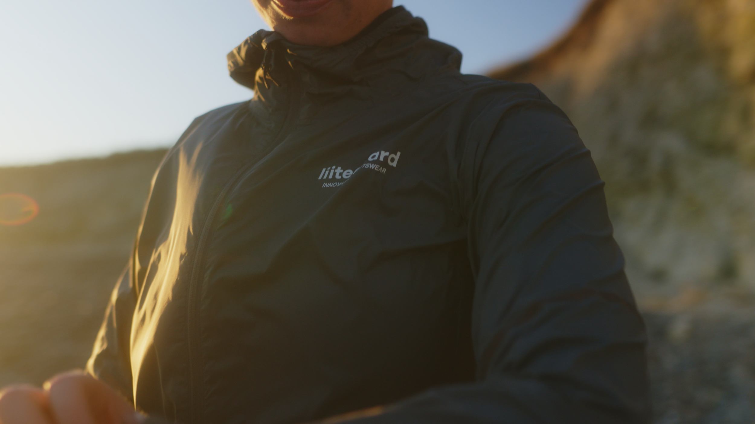 Close-up of a person wearing a black jacket with logos, with sunlight in the background, outdoors during sunset.