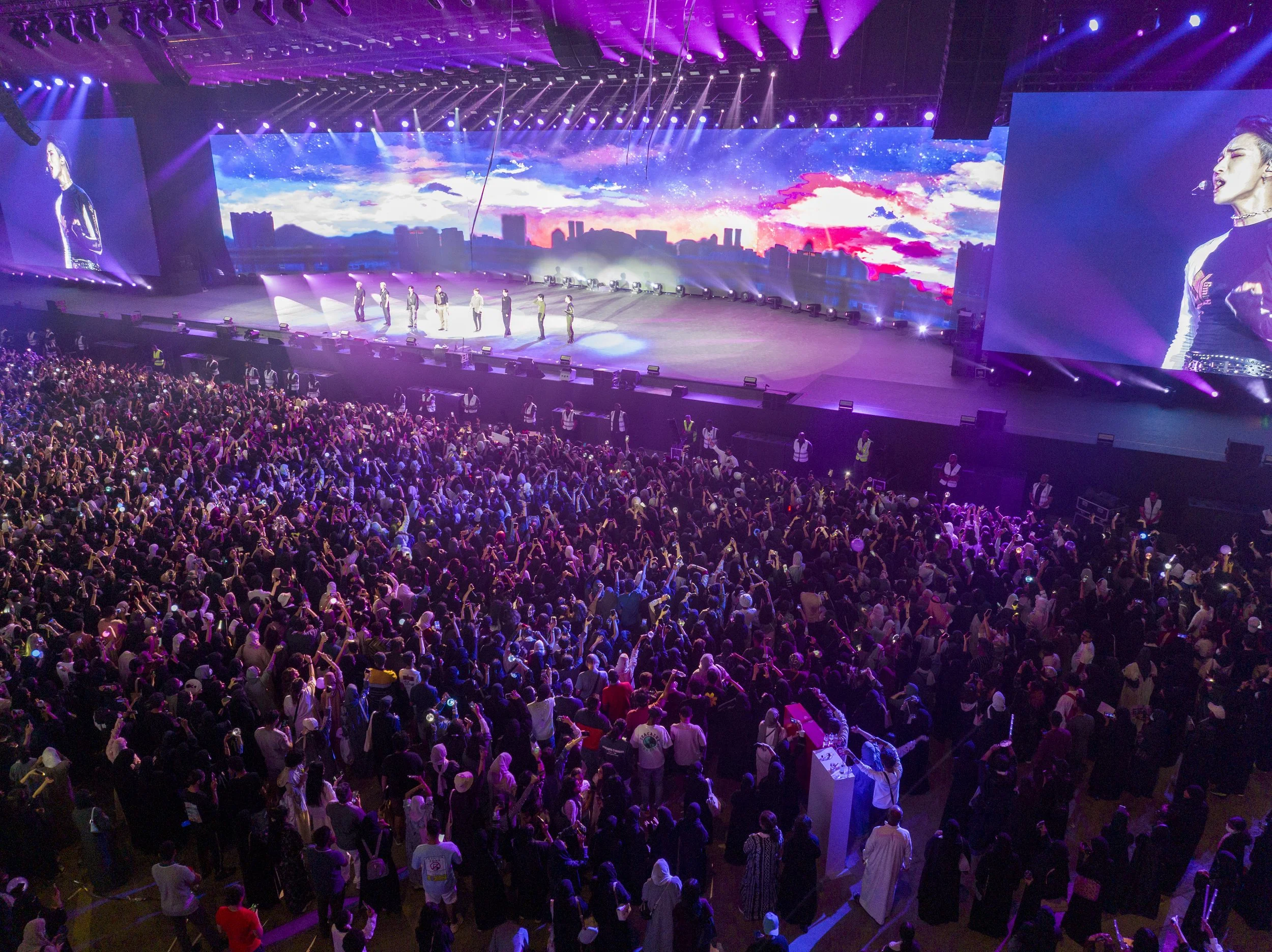 A large concert hall filled with a crowd of people watching a stage performance with bright purple and blue lighting, a digital city skyline backdrop on the screen, and performers on stage.