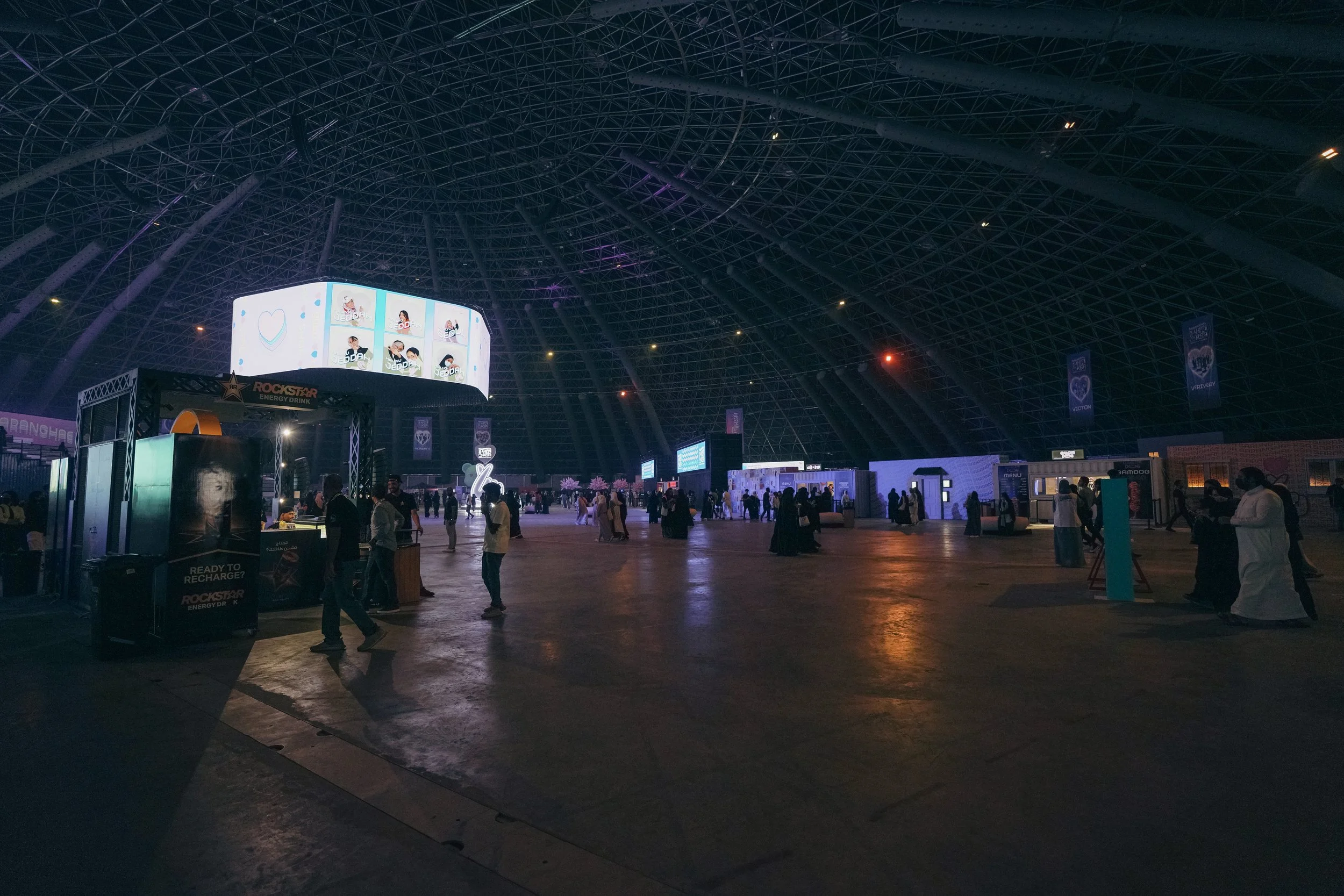 Interior of a large, dome-shaped event venue with people walking around, illuminated screens, promotional booths, and purple banners hanging from the ceiling.