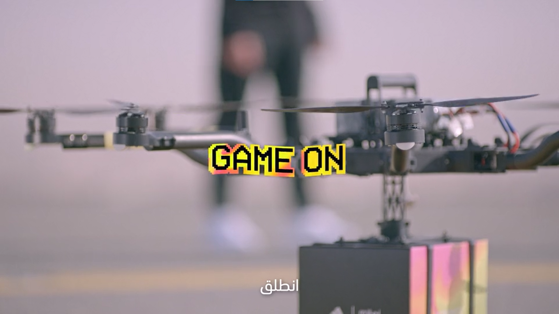 Close-up of a drone with the text 'GAME ON' in pixelated style overlayed, blurred person in the background, and small Arabic text at the bottom.