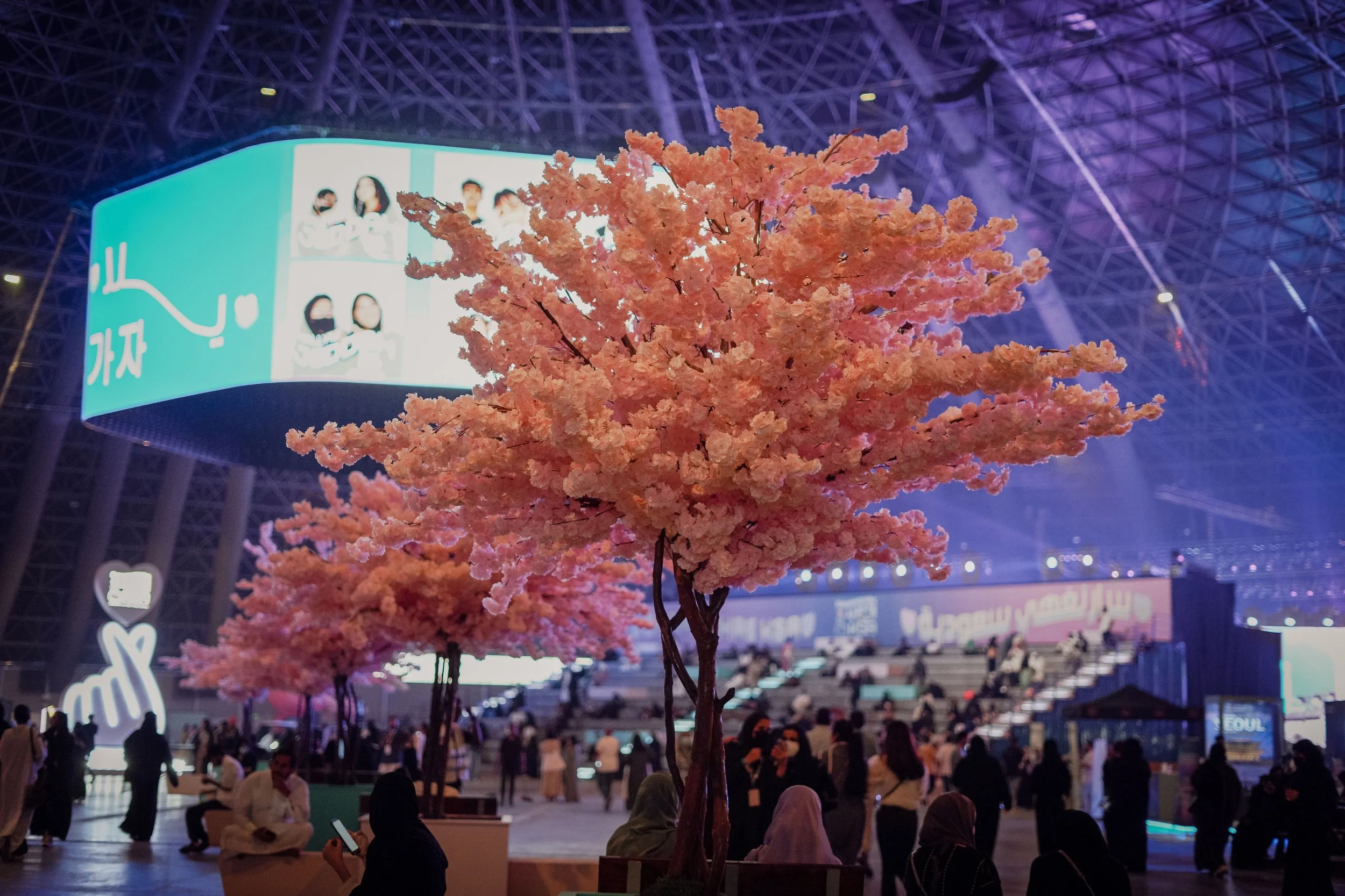 Pink artificial cherry blossom trees in an indoor venue with people sitting and standing, illuminated by purple and blue lights, with large screens and signage in the background.