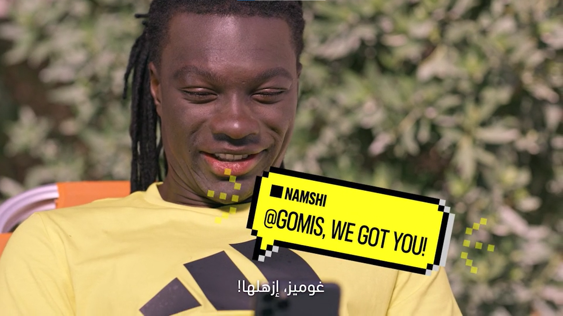 A man with dreadlocks wearing a yellow shirt is smiling while looking at a phone. There is a cartoon-style speech bubble with yellow background and black border in front of him that says '@Gomis, we got you!' and some digital effects. The background 