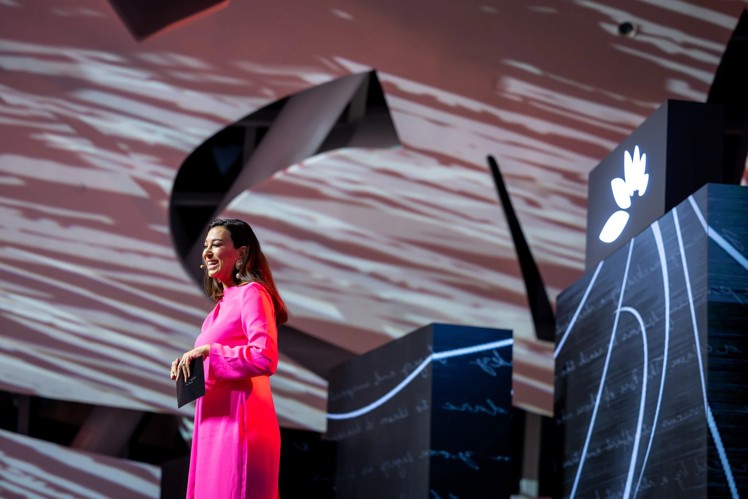 Woman wearing a bright pink dress standing on stage, smiling, holding a microphone, with decorative objects and a projected background in the scene.