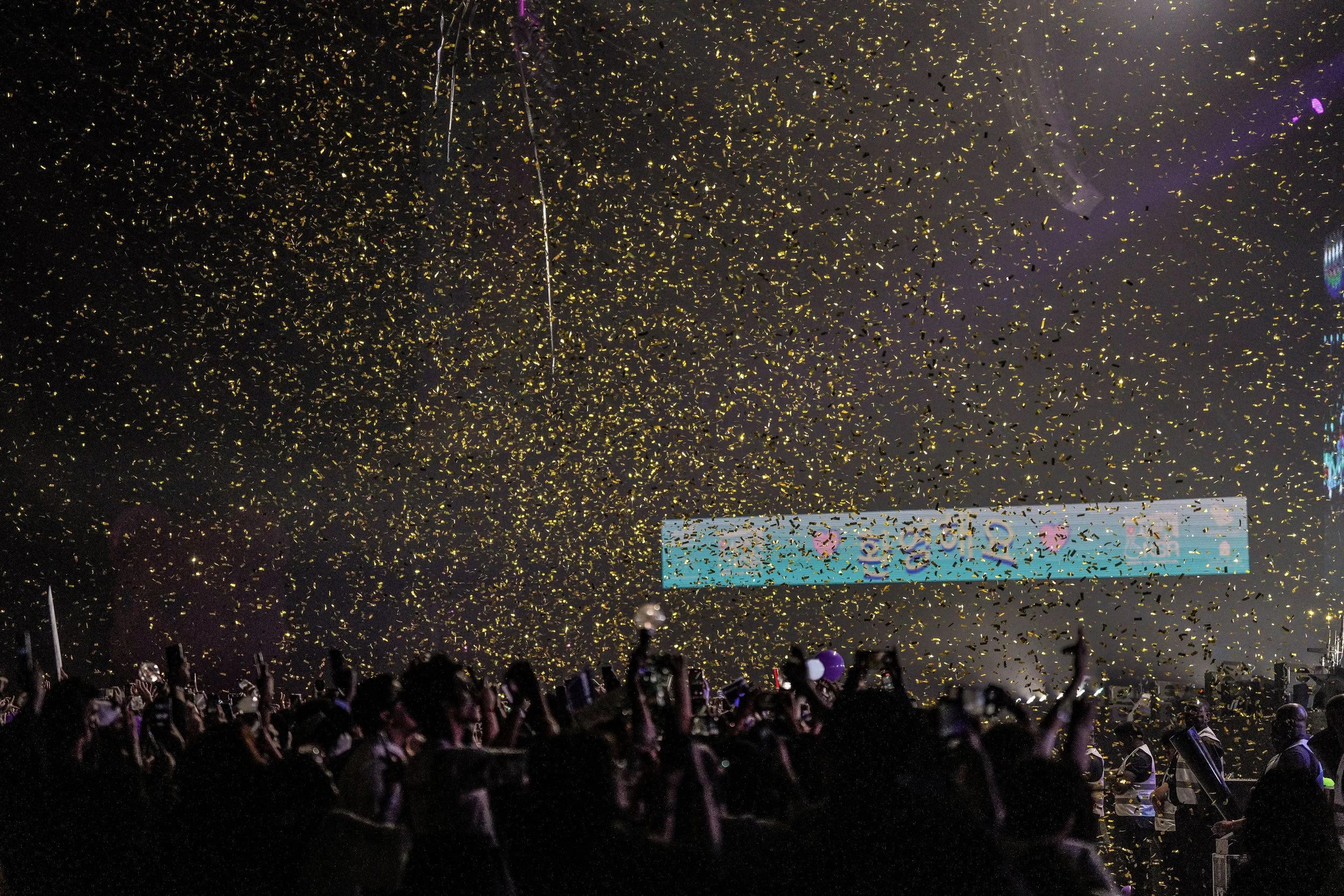 Crowd at an event with gold confetti falling from the ceiling, a large screen displaying colorful graphics in the background, and some people taking photos.